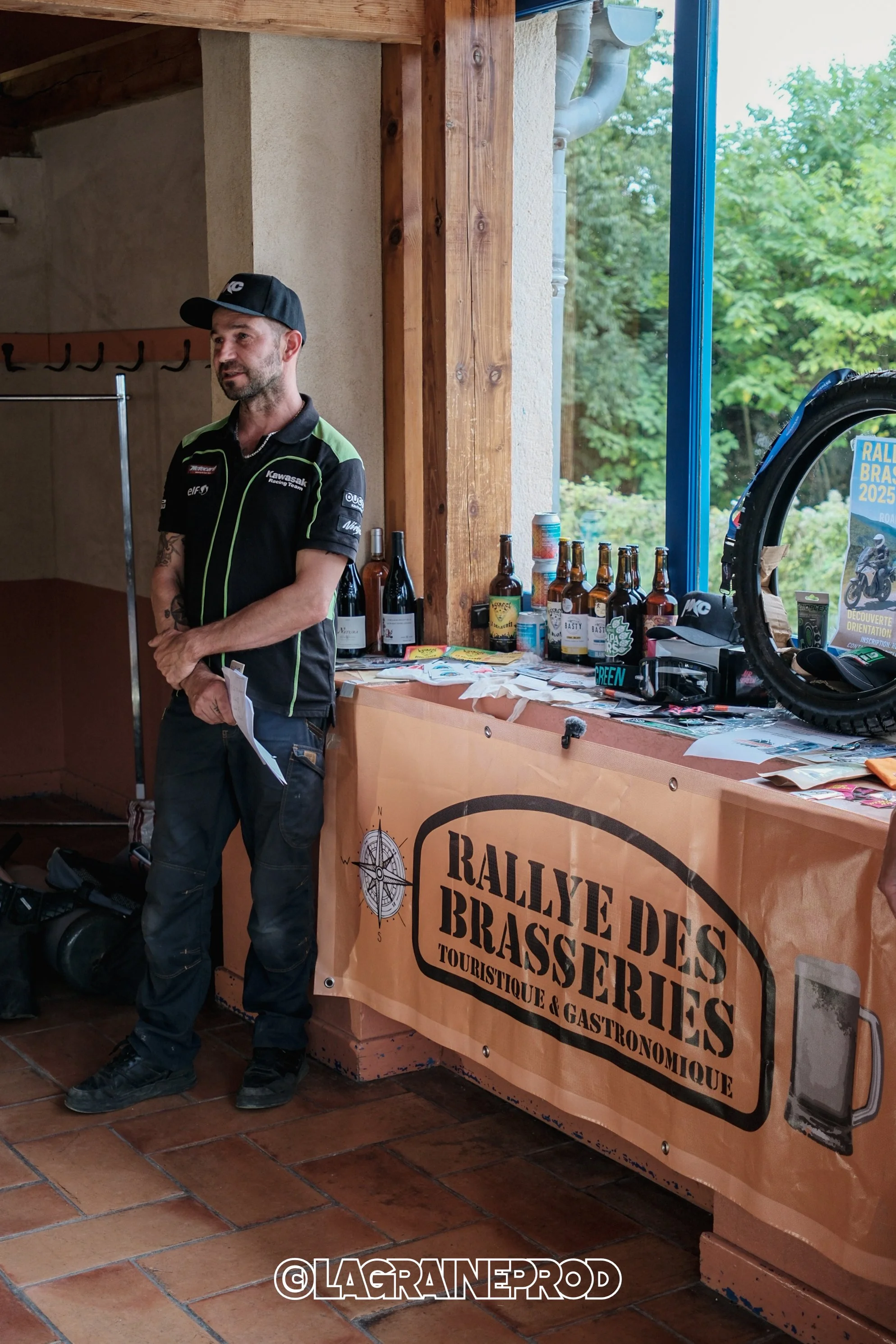 Un homme en tenue de pilote debout à côté d'une table avec un drapeau publicité pour le 'Rallye des Brasseries', entouré de bouteilles de bière, de flyers et d'autres objets promotionnels; à l'arrière-plan, vue sur la nature par une fenêtre.