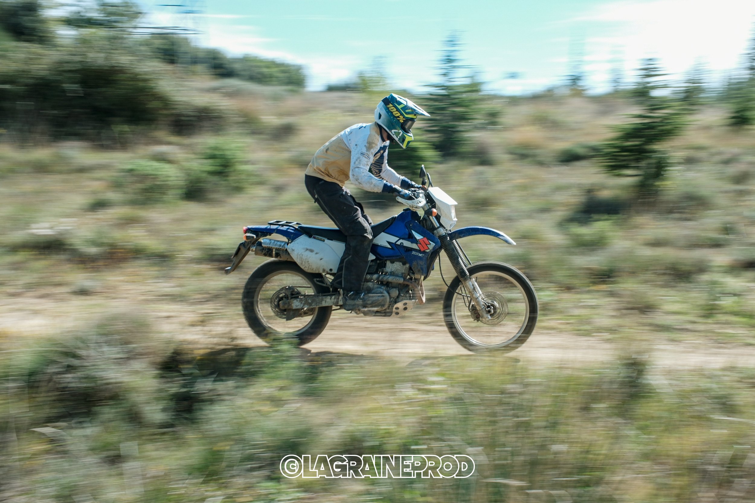 Un motocycliste portant un casque et des vêtements de protection, chevauchant une moto tout-terrain sur un chemin rural avec de la végétation en arrière-plan.