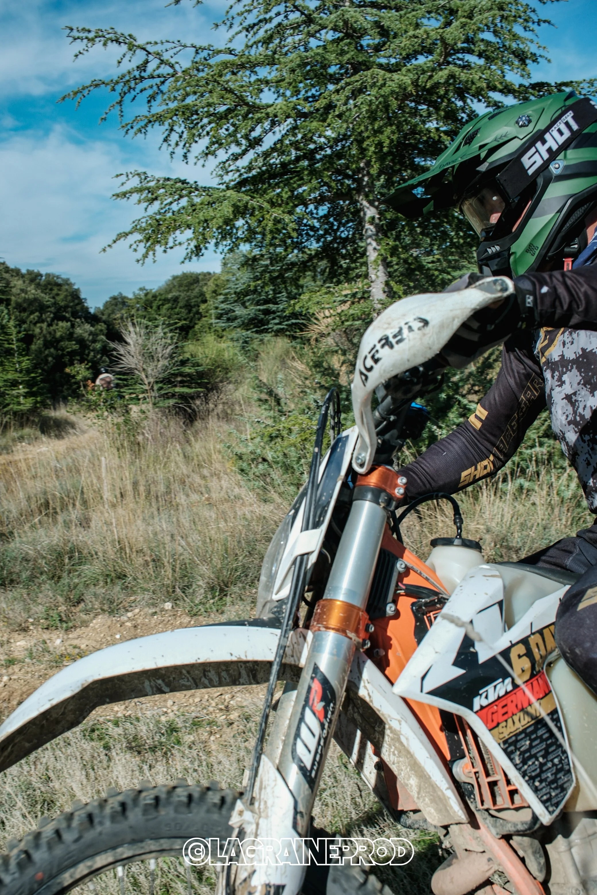 Un motard portant un casque vert et des vêtements de protection, chevauchant une moto tout-terrain orange et blanc sur un chemin de campagne, avec des arbres et un ciel bleu en arrière-plan.