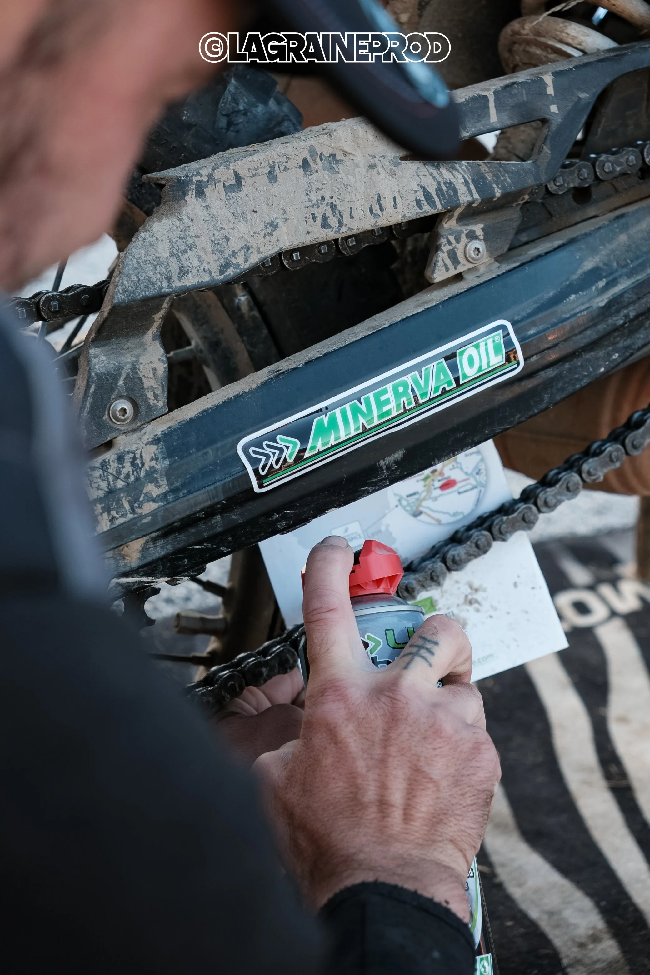 Une personne applique de la lubrification sur la chaîne d'une moto tout-terrain, avec un autocollant Minerva Oil visible sur le cadre et une carte ou un plan posé en dessous. La scène se déroule en extérieur, probablement lors d'une activité de motoc