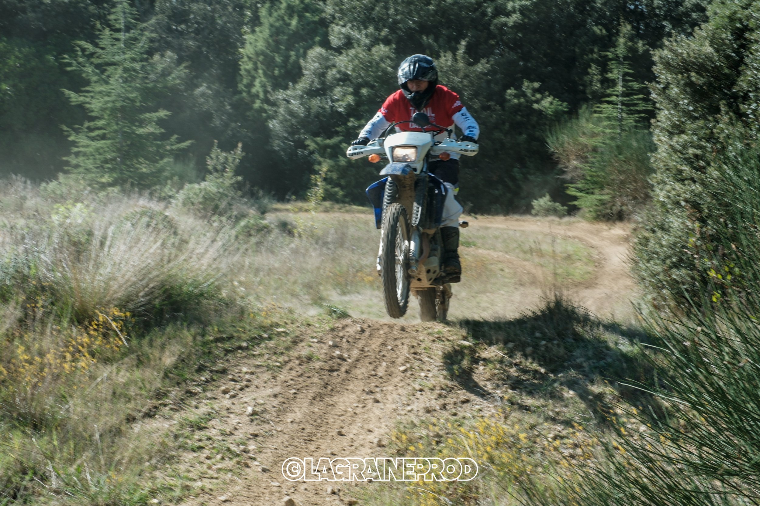 Une personne portant un casque et des vêtements de protection, faisant du motocross sur un sentier en pleine nature, avec des arbres en arrière-plan.