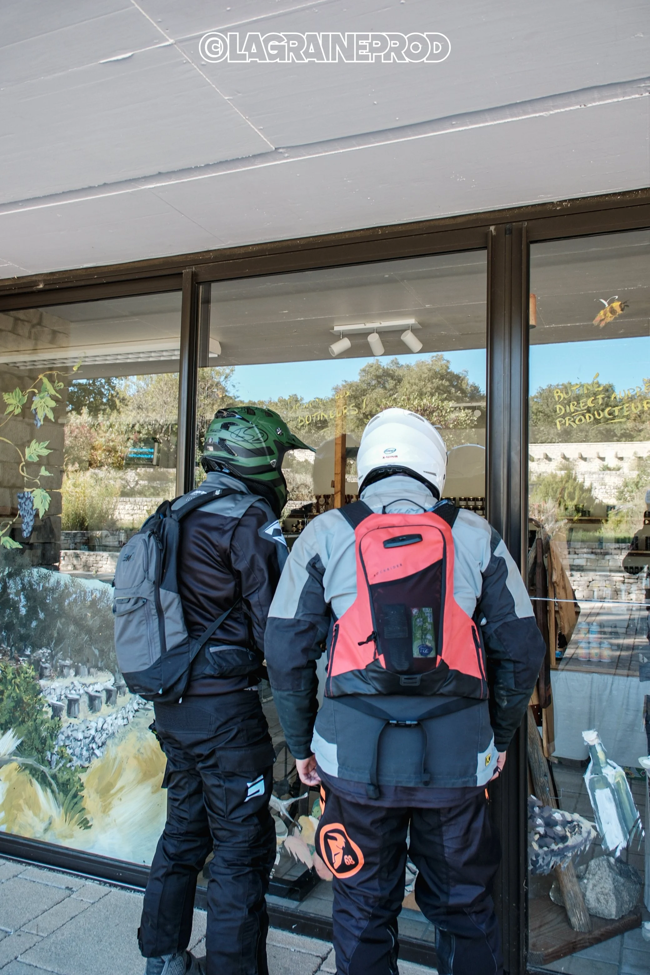 Deux motards regardant à travers une vitrine, portant des casques et des sacs à dos, à l'extérieur d'un magasin.