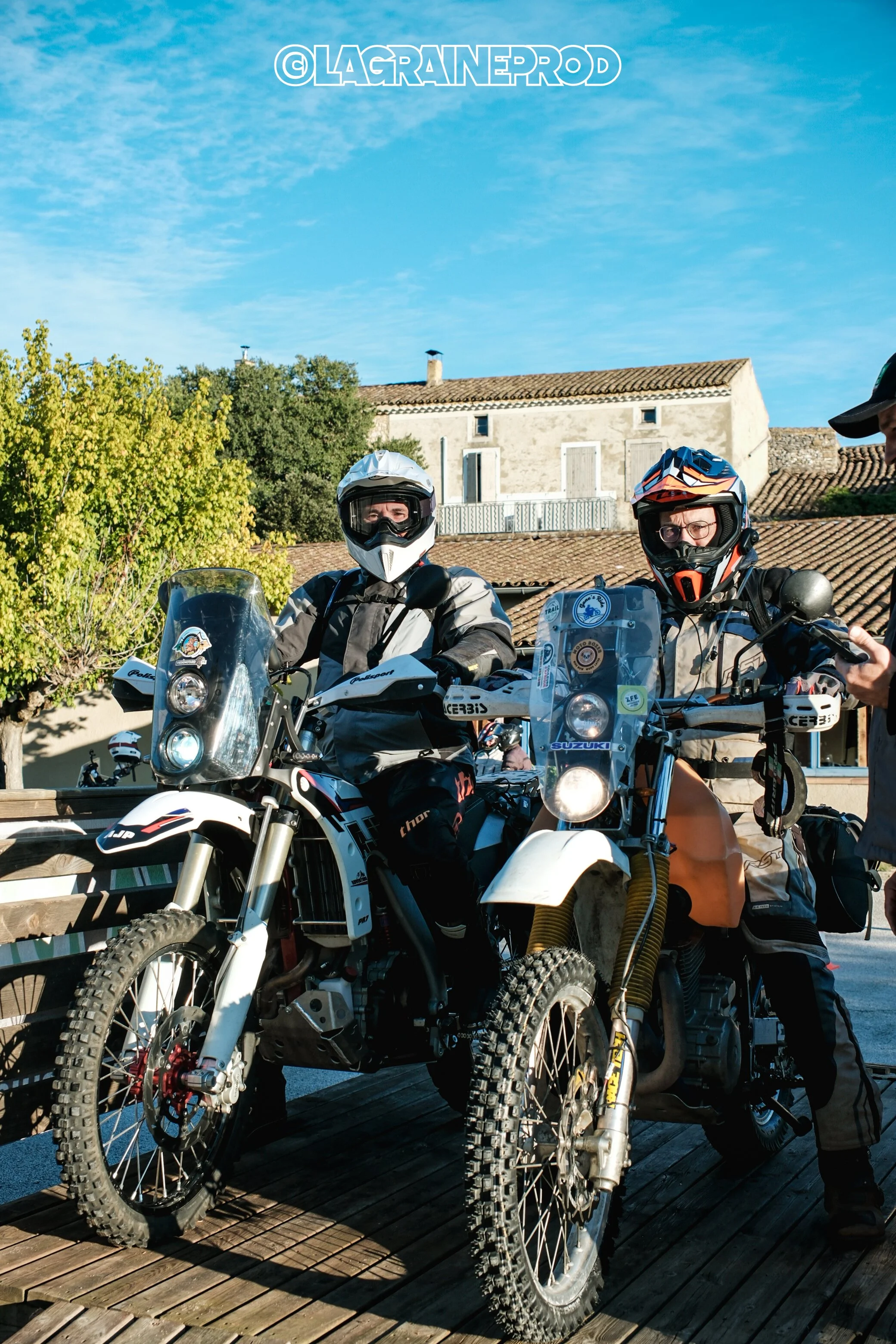 Deux motocyclistes en tenue de moto et casque prêts pour une aventure, sur des motos tout-terrain, à l'extérieur avec un ciel bleu et des maisons en arrière-plan.