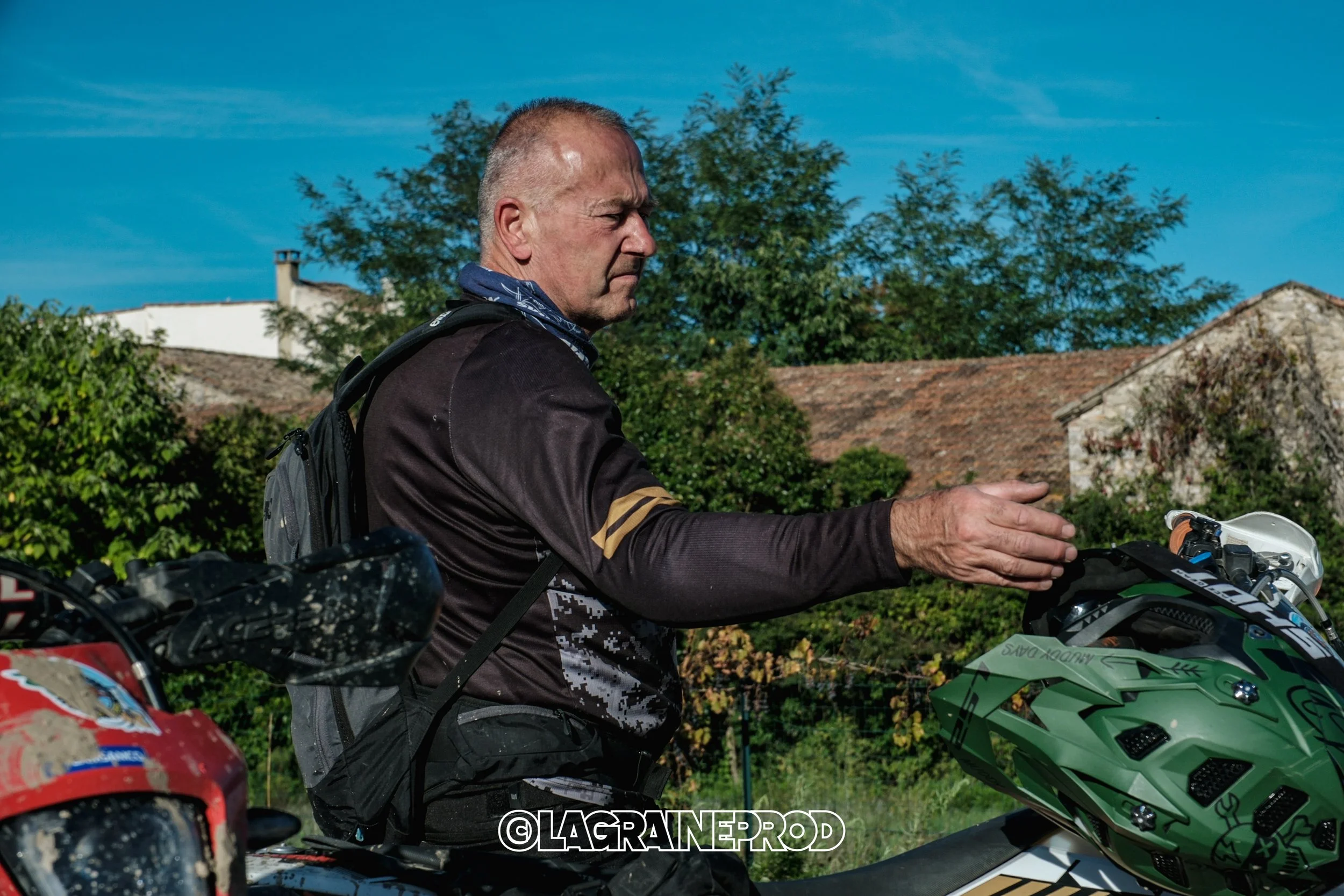 Un homme en vêtements de moto ajustant son casque vert sur un moteur de moto, avec un fond de maisons et d'arbres sous un ciel bleu.