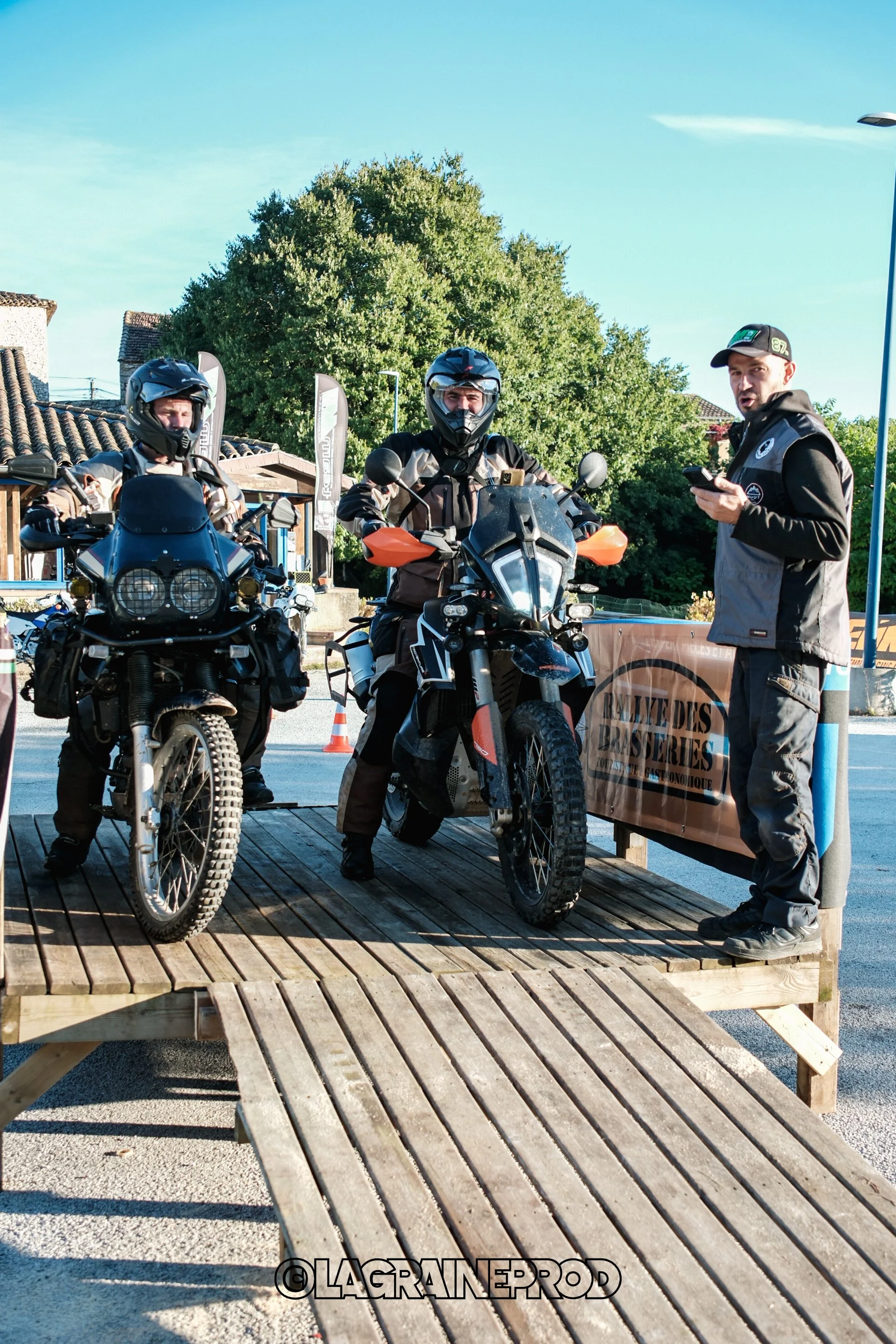 Trois motards portant des équipements de protection en train de discuter lors d'un événement en plein air, avec une plateforme en bois et un panneau publicitaire.»}]]}]{];]  }  ));}  сереб} (json-schema-from-ImageAltText)  @{}]}7/}  |}  }  Öff(ent)}]