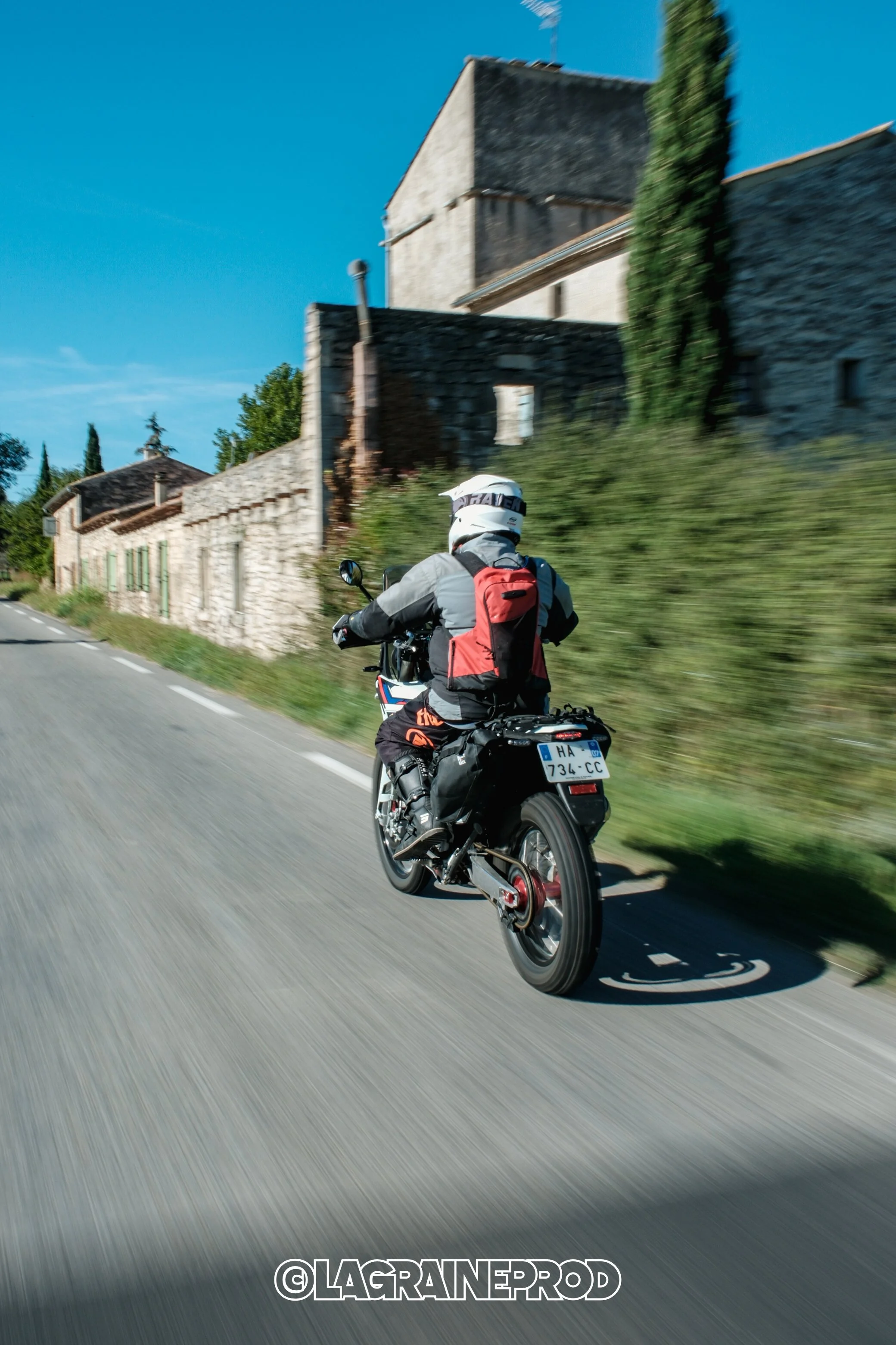 Motard voyageant sur une route de campagne en France, avec maisons anciennes en pierre et cypress en arrière-plan, ciel bleu clair.