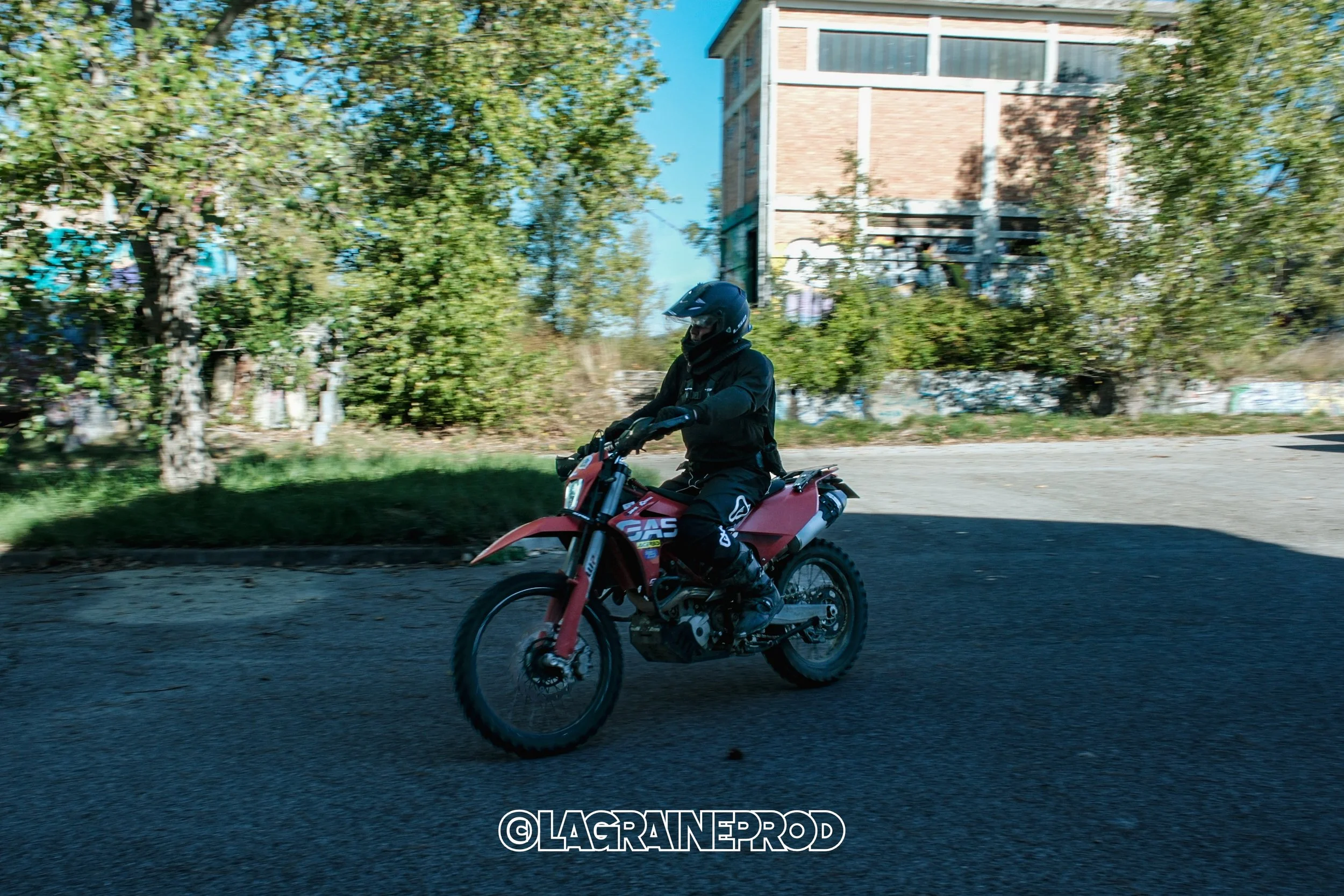 Personne en tenue noire portant un casque, roulant à moto rouge sur une route en extérieur avec arbres et bâtiment en arrière-plan.