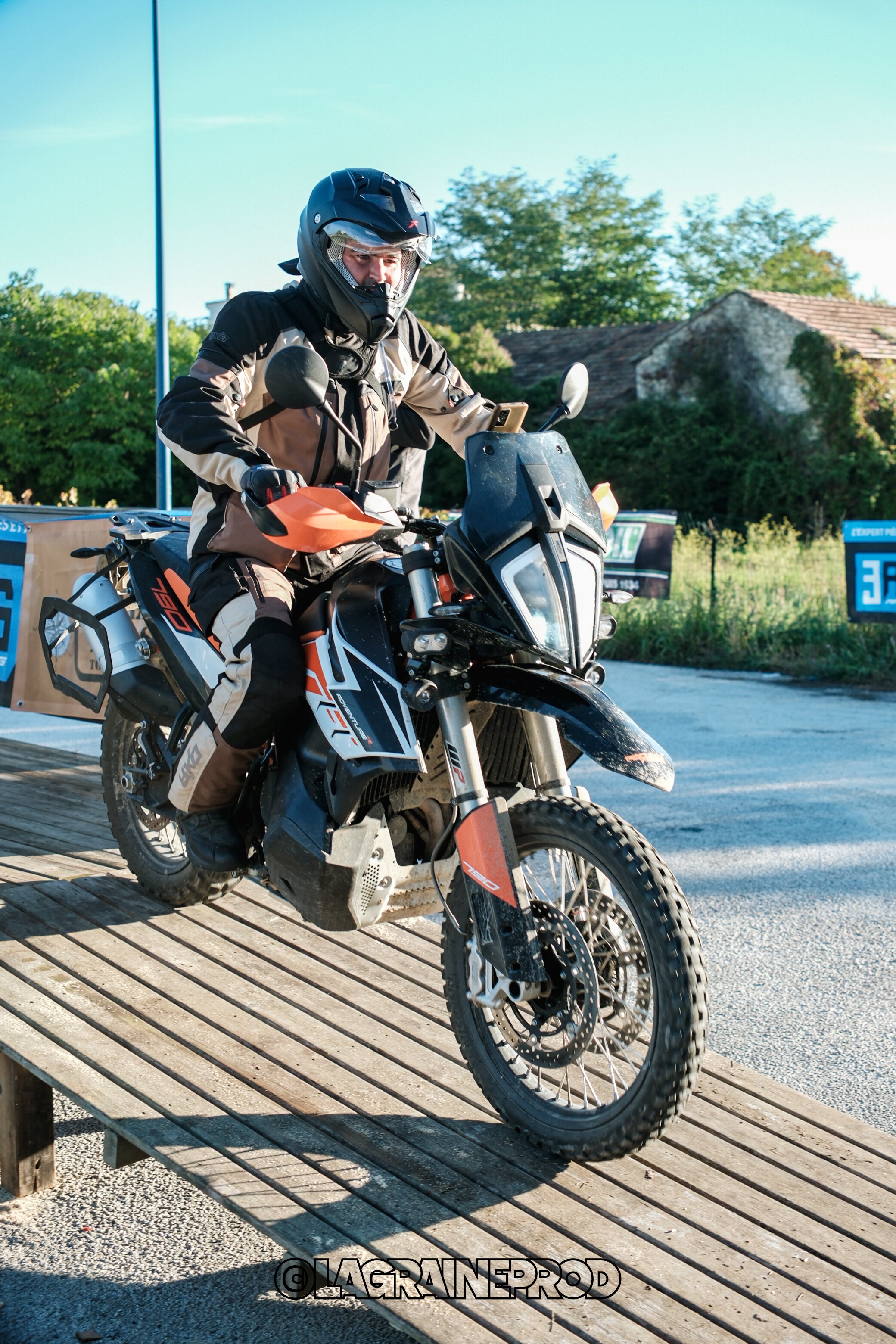 Un homme en équipement de moto tout-terrain sur une moto noire et orange, prêt à partir, en dehors d'une maison en pierre, avec des arbres en arrière-plan.