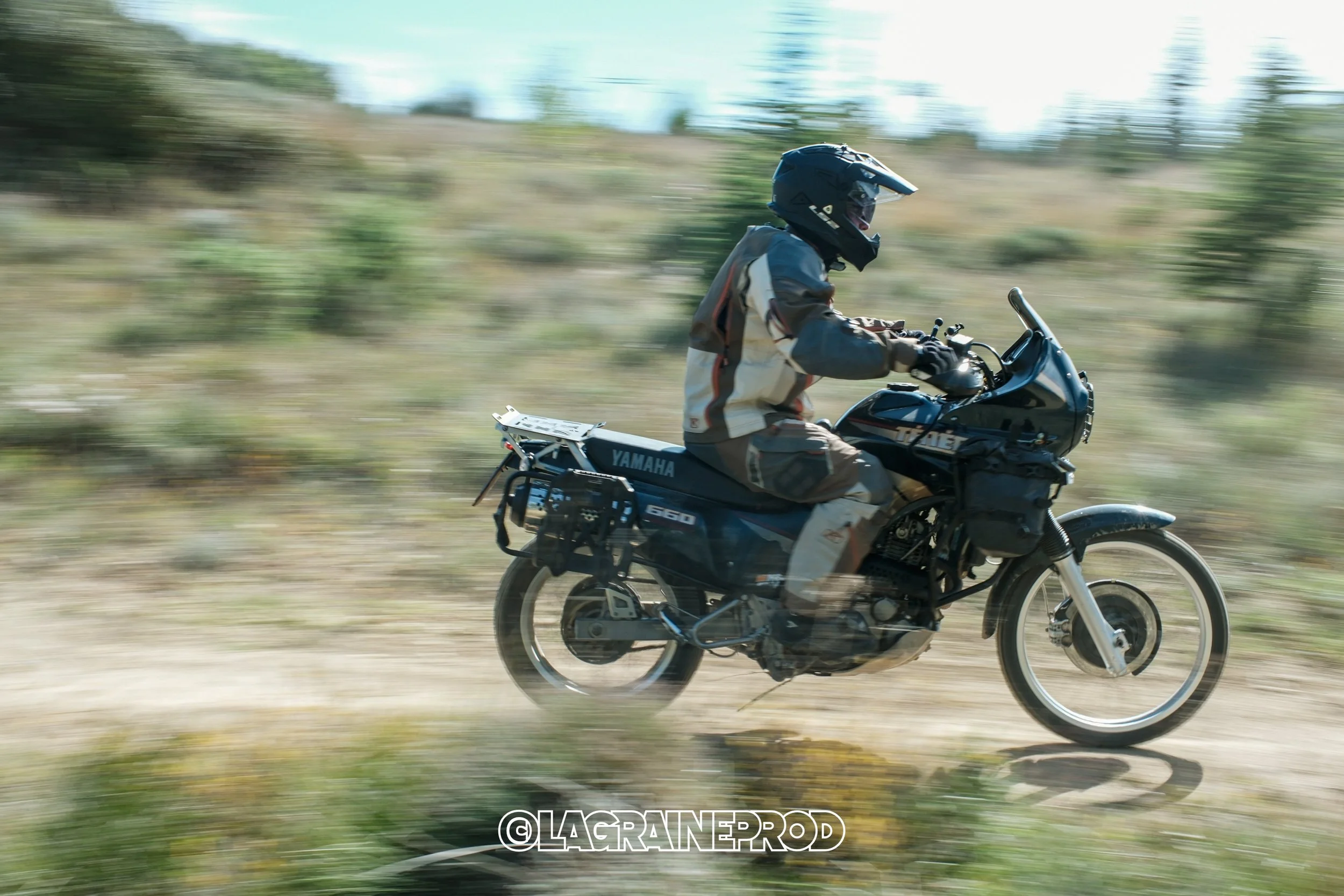 Un motard portant un casque et une veste de motocross, roulant à vive allure sur une moto Yamaha dans un paysage naturel ouvert avec de la végétation. La photo montre un flou de mouvement, suggérant la vitesse.