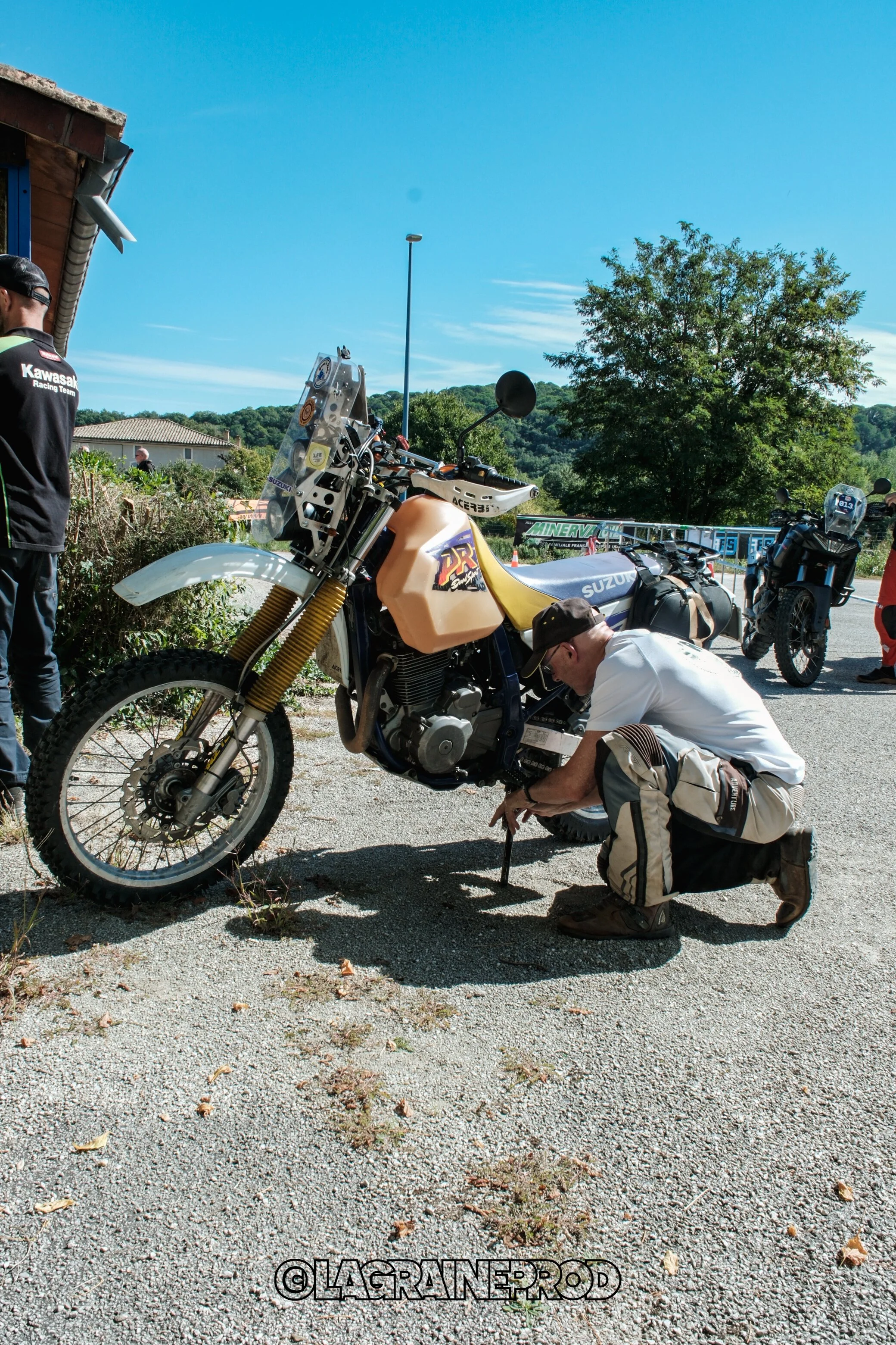 Homme en train de réparer une moto Suzuki en plein air sous un ciel bleu, avec d'autres motos en arrière-plan.
