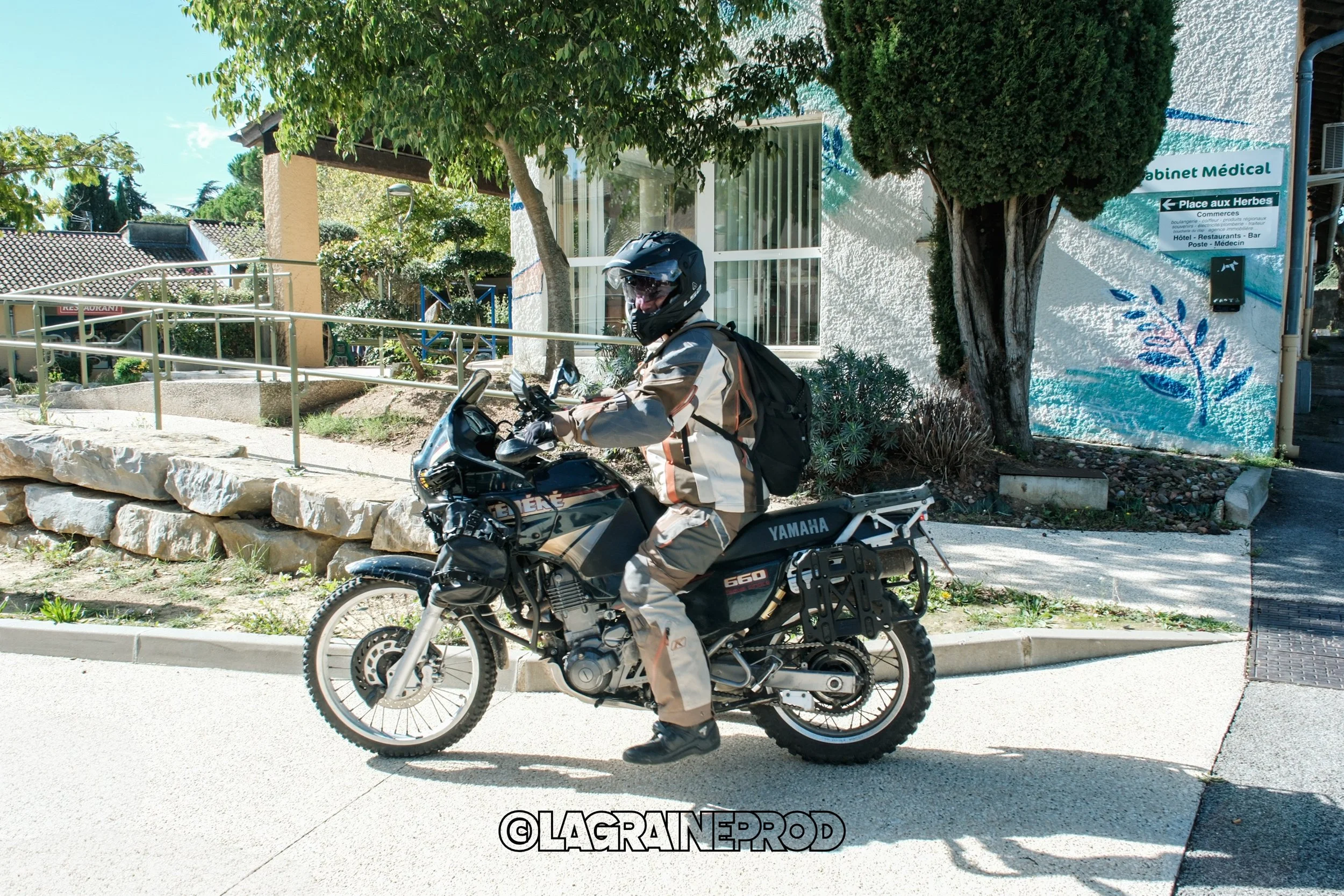 Motard en tenue de protection et casque, assis sur une moto Yamaha noire, à l'extérieur d'un bâtiment avec des arbres et des panneaux en arrière-plan.