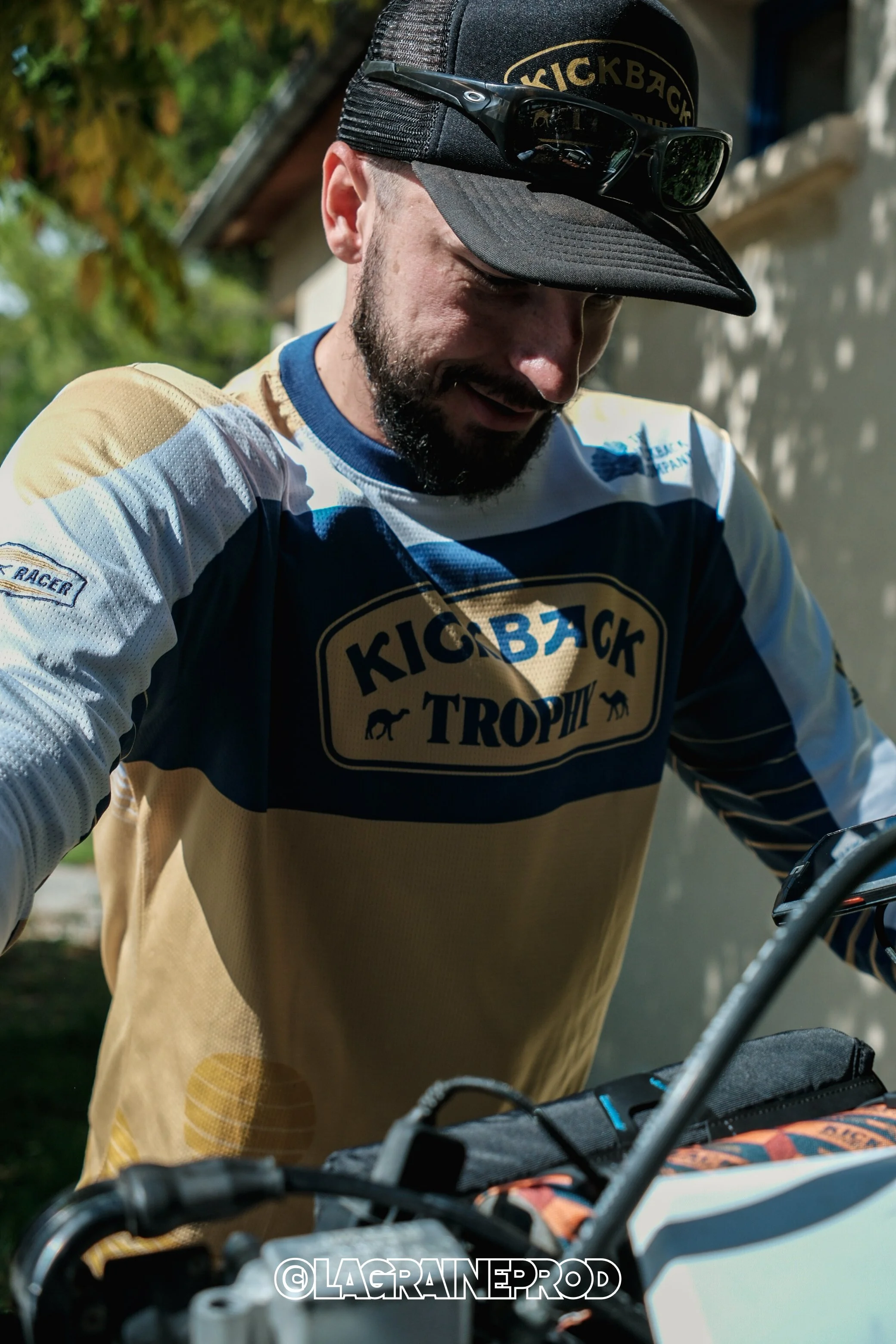 Un homme portant un maillot de cyclisme avec le logo 'Kick Back Trophy' et une casquette noire avec des lunettes de soleil posées dessus, se préparant pour une activité de motocross ou de vélo tout terrain.