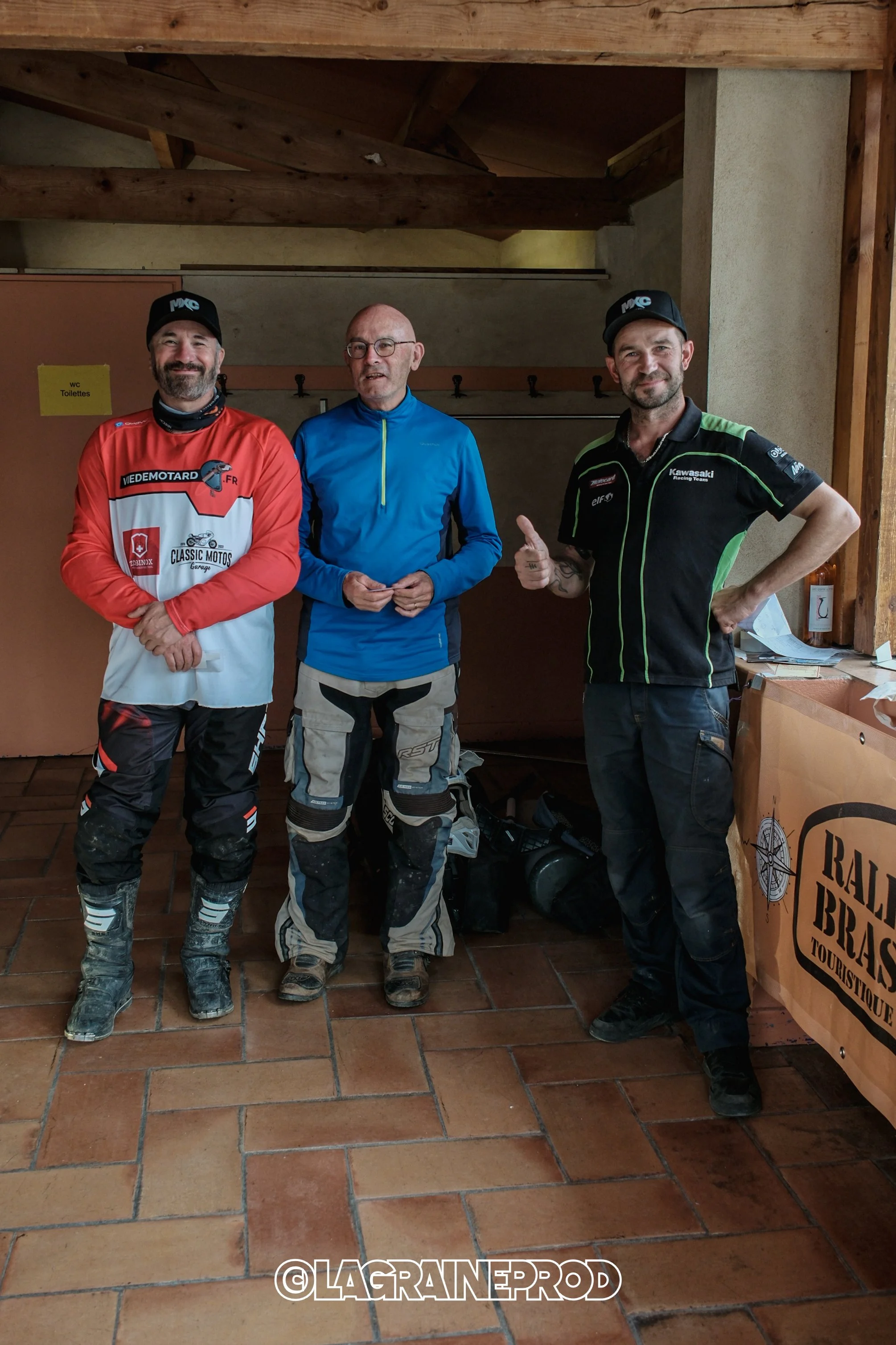 Trois hommes en vêtements de motocross se tiennent dans une pièce avec un sol en carreaux et un panneau derrière eux. L'un des hommes fait un signe de pouce levé.