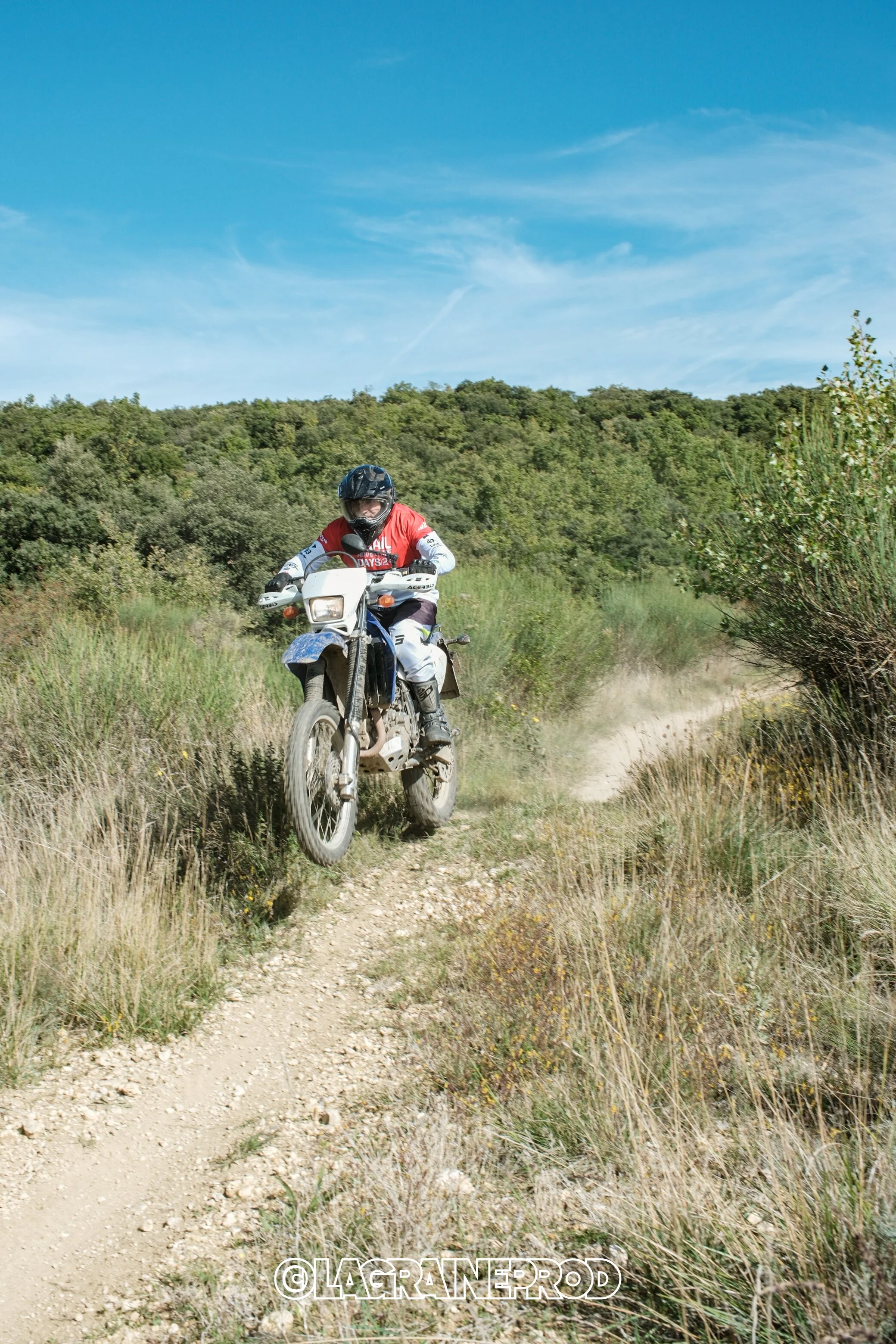 Un homme chevauchant une moto tout-terrain sur un sentier de terre dans un paysage naturel avec des buissons et des arbres, sous un ciel bleu clair.