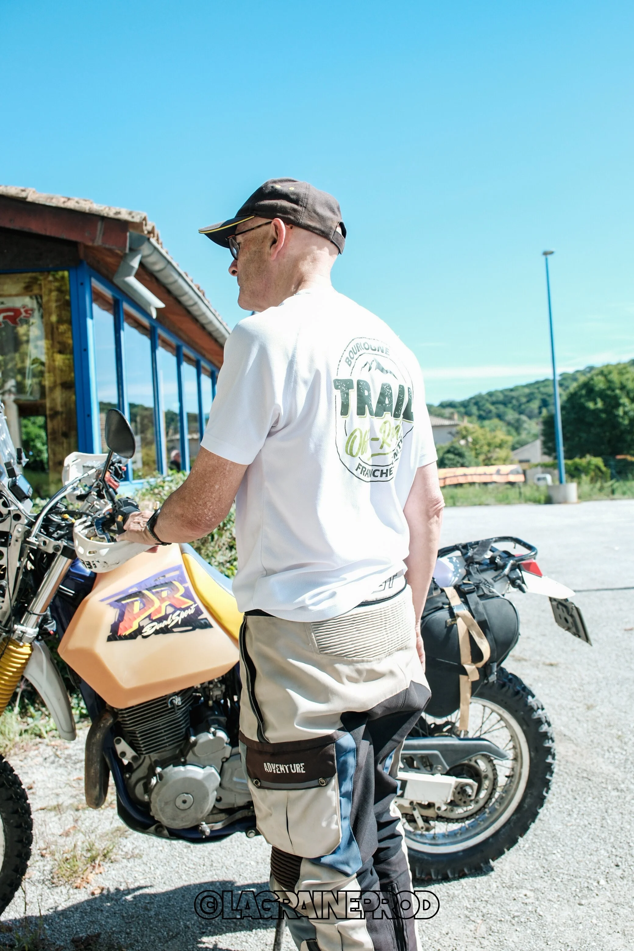 Un homme avec un casque de moto, portant une chemise blanche et un pantalon de moto, se tenant à côté d'une moto tout-terrain dans un paysage en plein air avec un ciel bleu clair.