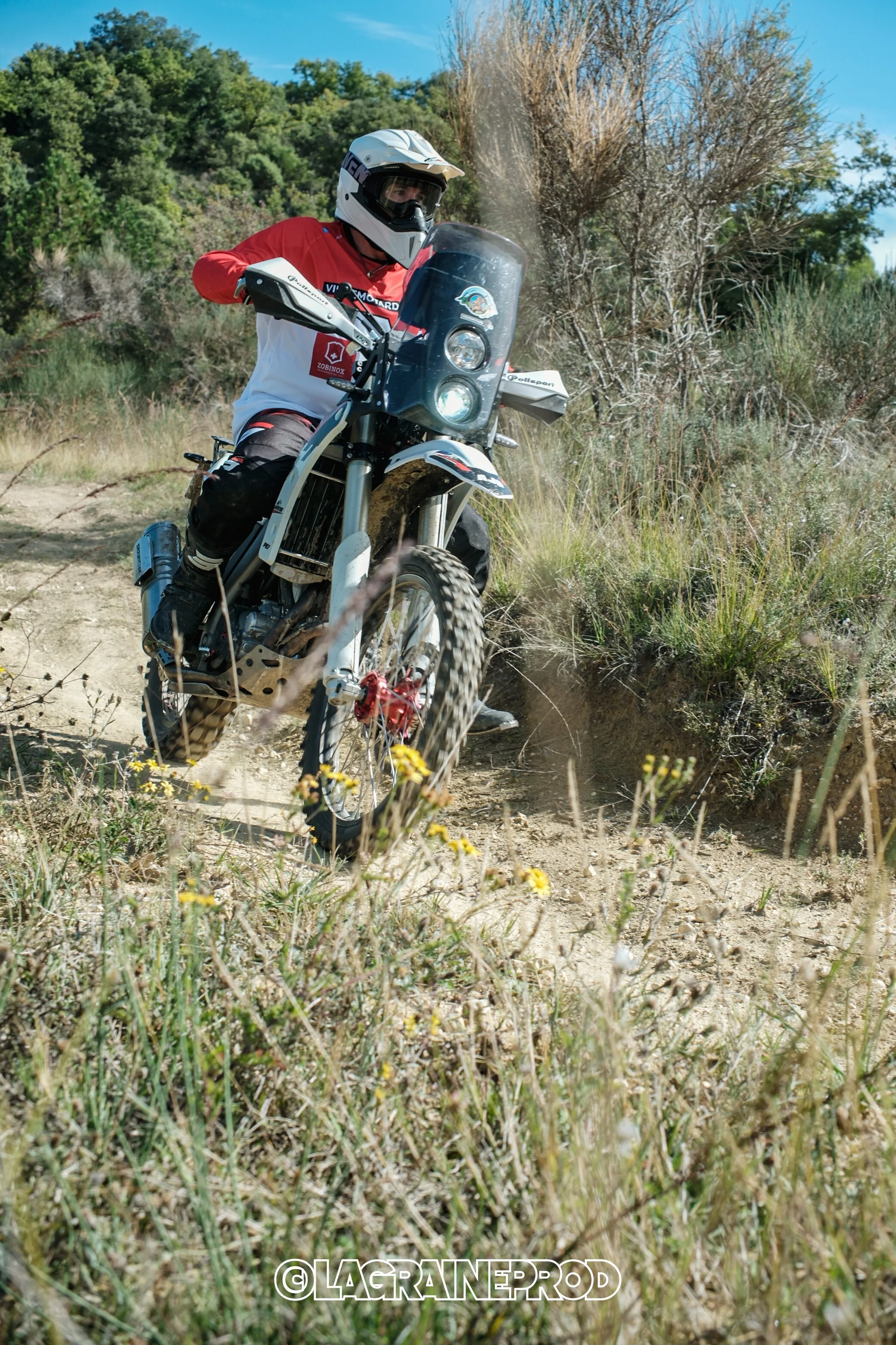 Un motard portant un casque blanc et une veste rouge et blanche, roulant à moto tout-terrain sur un sentier en nature avec des arbres et de la végétation sèche.