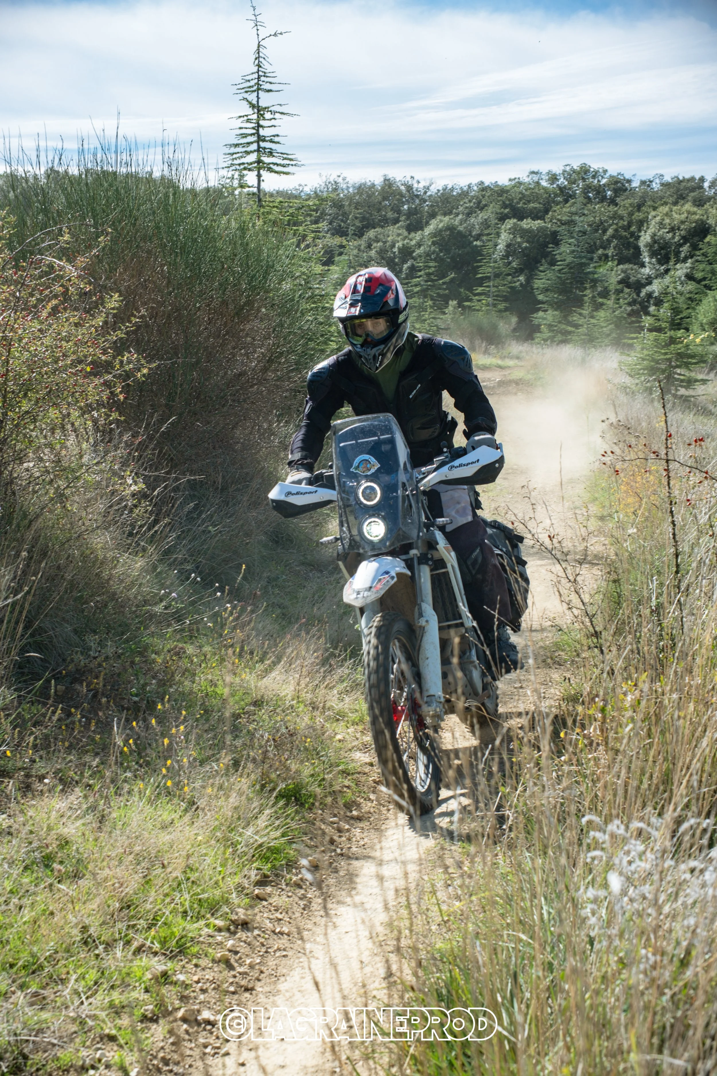 Un motard en tenue de protection, portant un casque, fait de la moto tout-terrain sur un sentier en pleine nature avec des arbustes et des arbres autour.