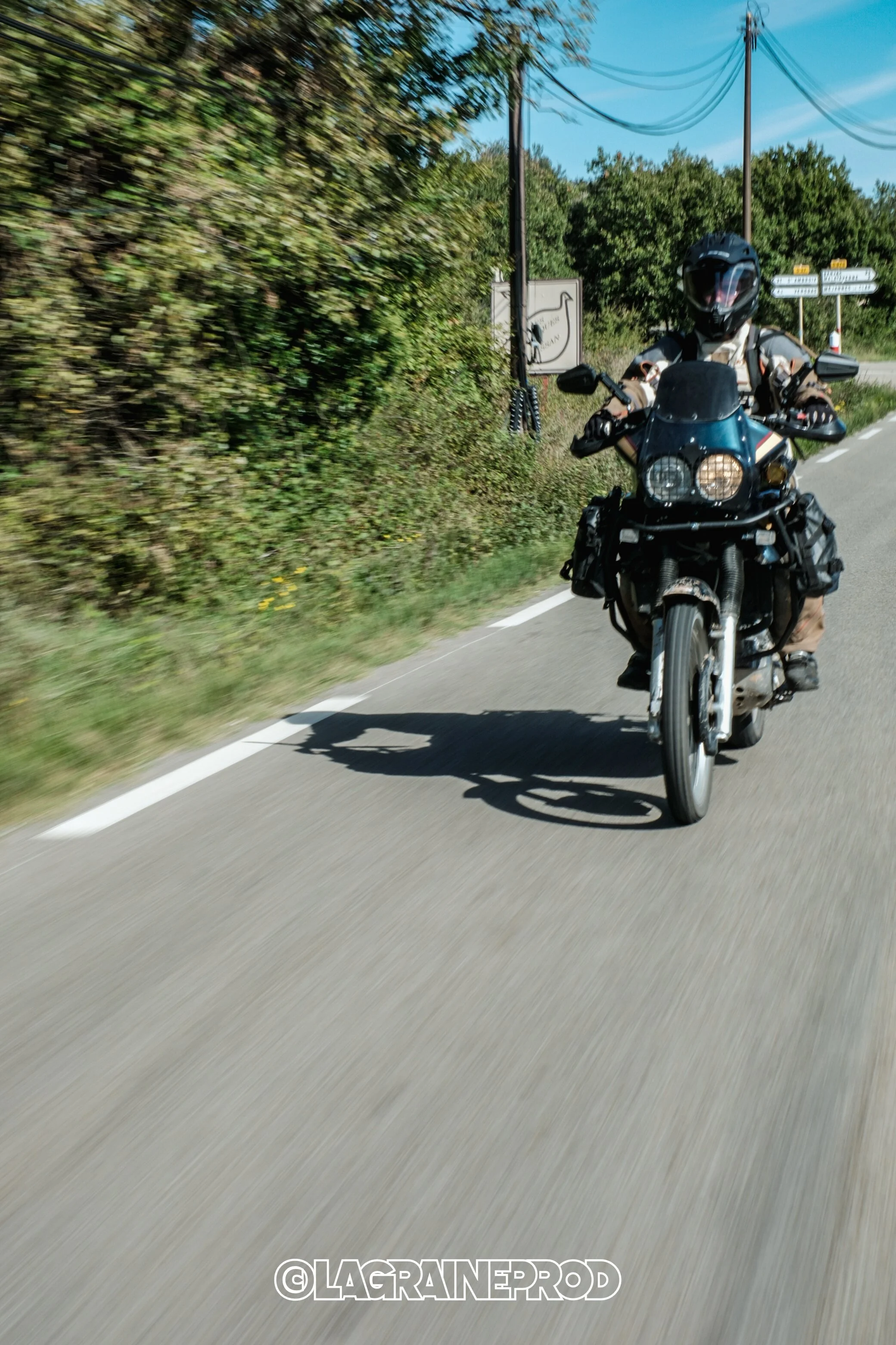 Un motard portant un casque noir et une veste de moto, roulant à moto sur une route en zone rurale avec des arbres et un panneau de signalisation en arrière-plan.