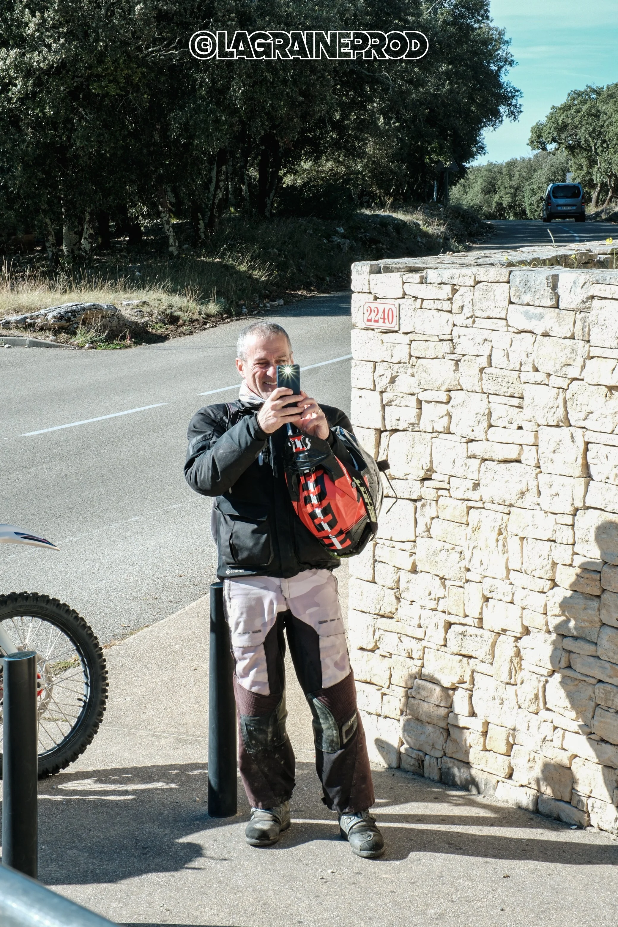 Un homme souriant en tenue de moto prend une photo avec son téléphone devant un mur en pierre, sur une route en campagne.