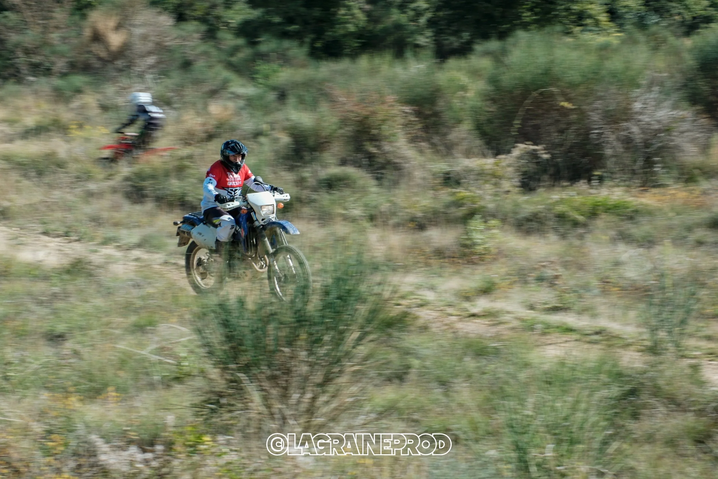 Deux motocyclistes roulant à travers un terrain herbeux, l'un en avant de l'autre, dans un environnement naturel avec des buissons et des arbres