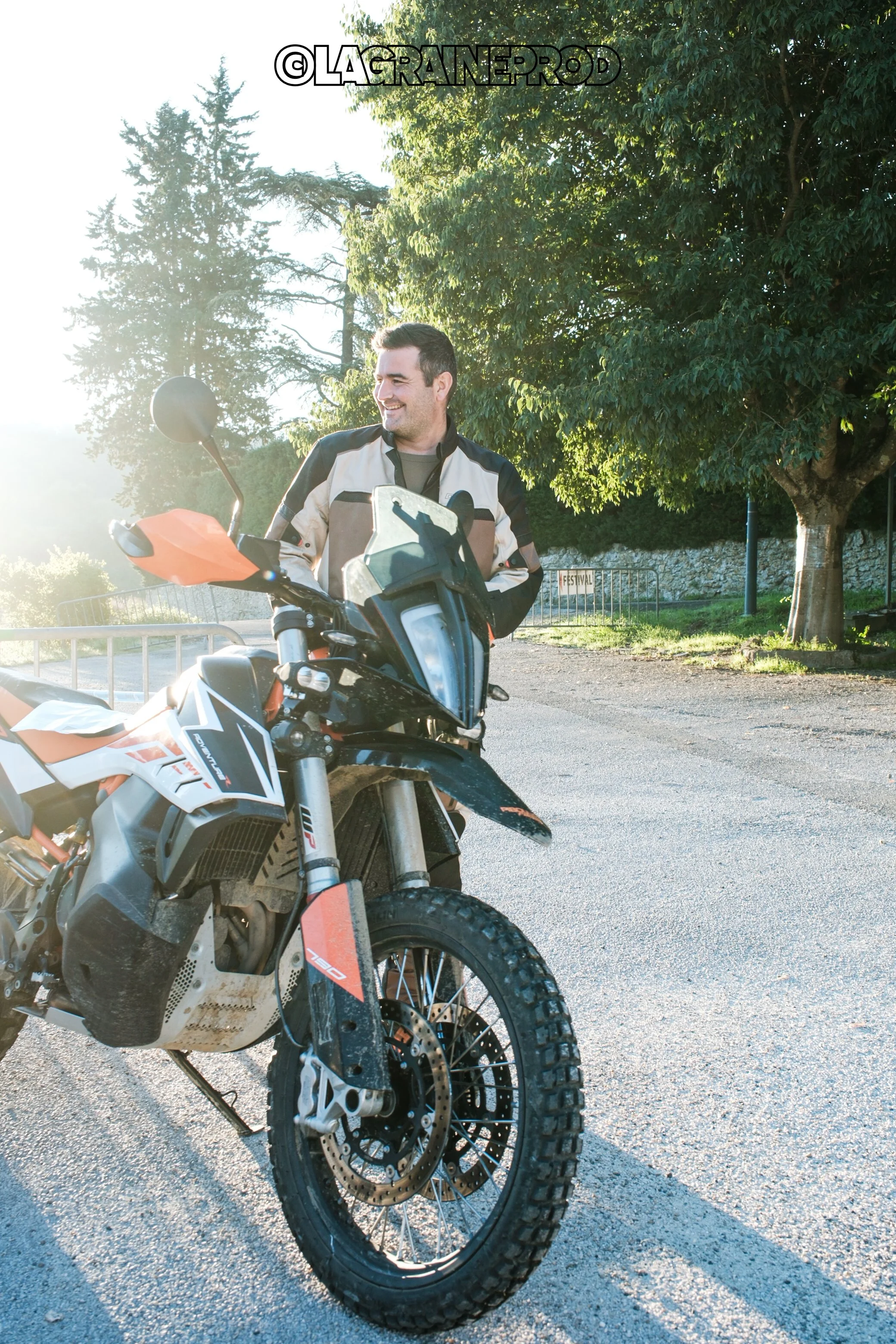 Un homme souriant avec une veste de moto se tient à côté d'une moto tout-terrain sur une route ensoleillée, avec des arbres en arrière-plan.