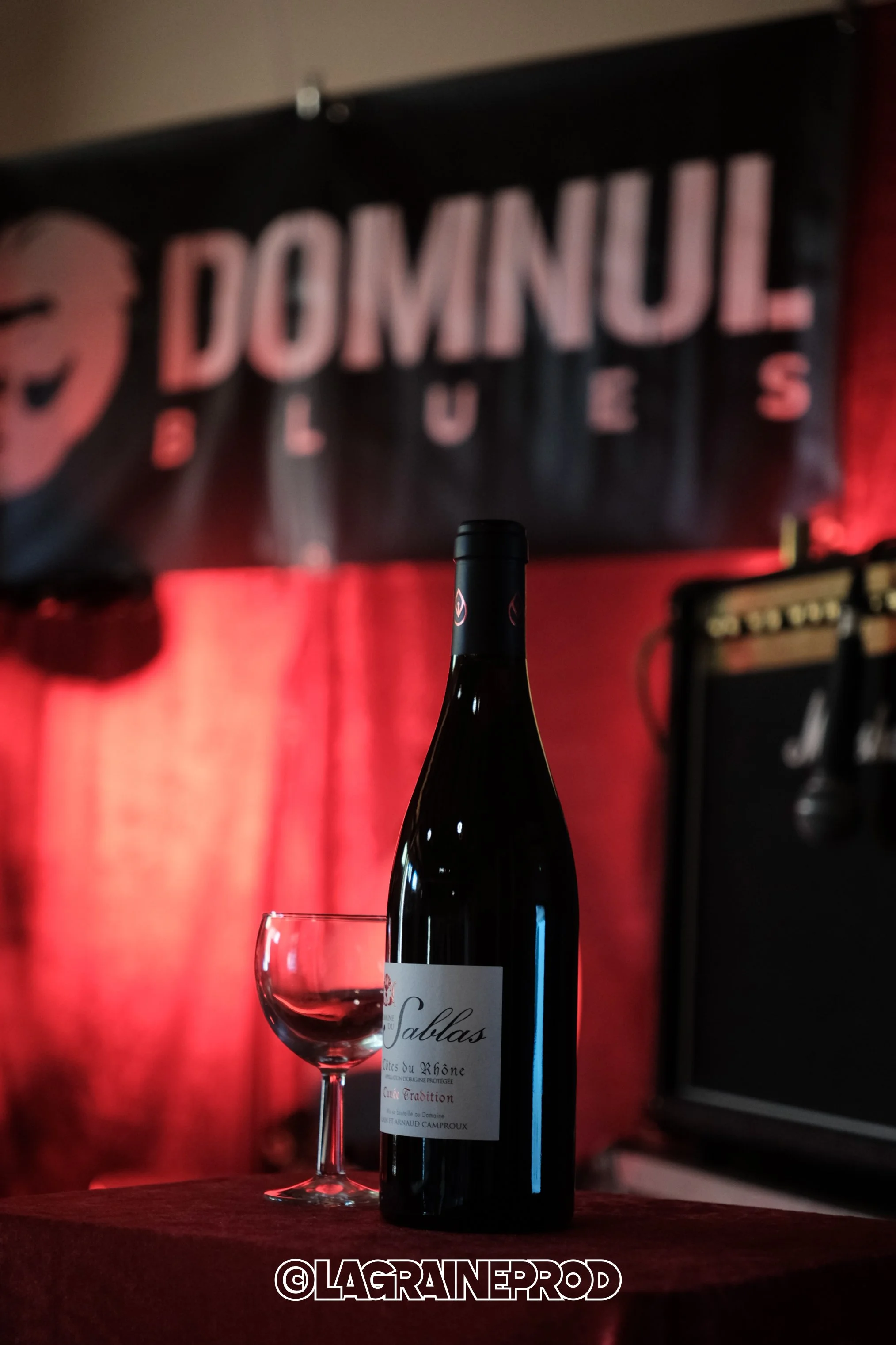 Une bouteille de vin rouge et un verre à vin vide sur une table en tissu rouge. En arrière-plan, un panneau noir avec l'inscription déformée "DOMINUL" en lettres roses. L'ambiance est tamisée avec des lumières rouges.