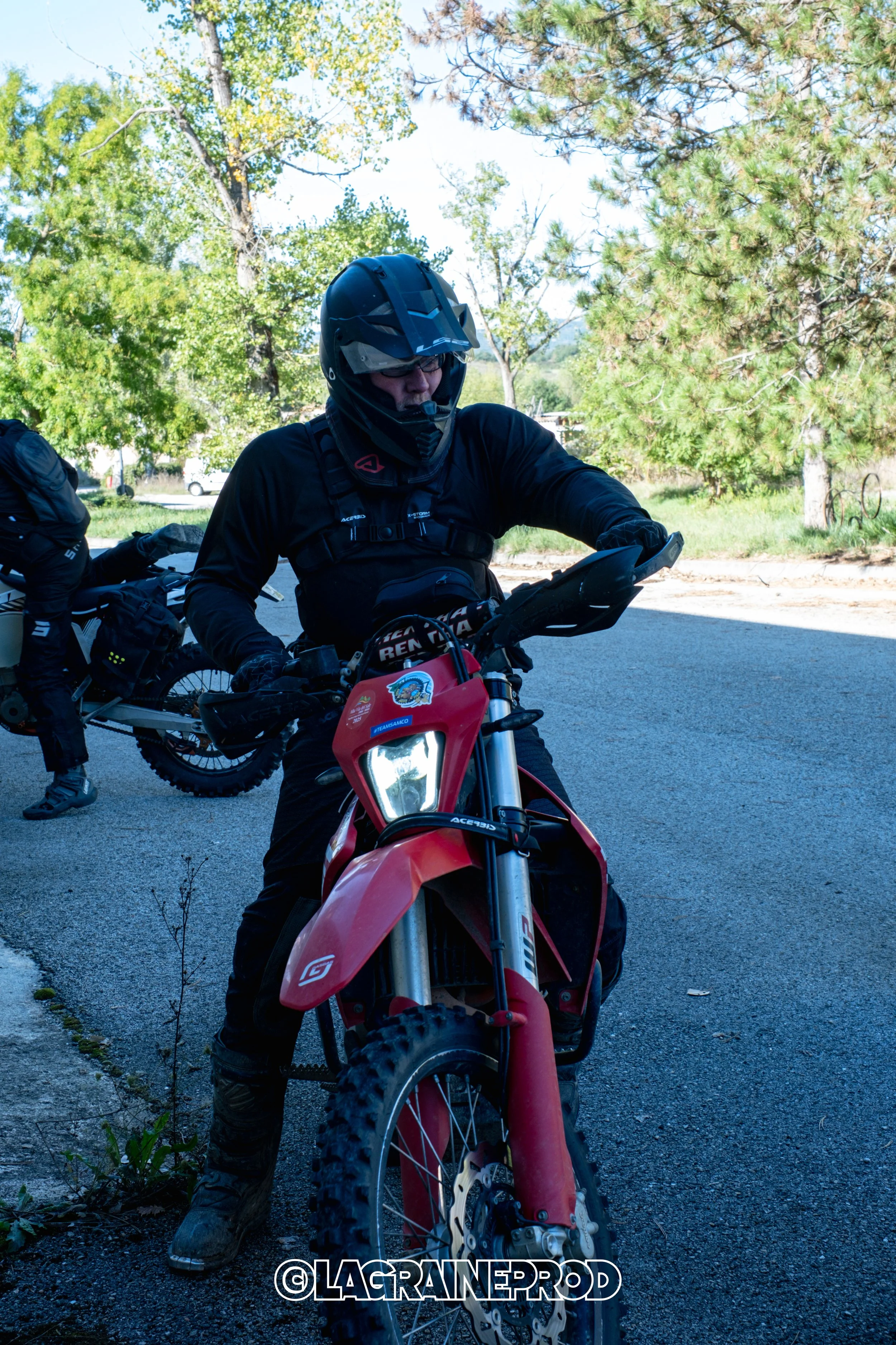 Personne portant un casque et des vêtements de moto tout-terrain, assise sur une moto rouge, en plein air avec des arbres en arrière-plan.