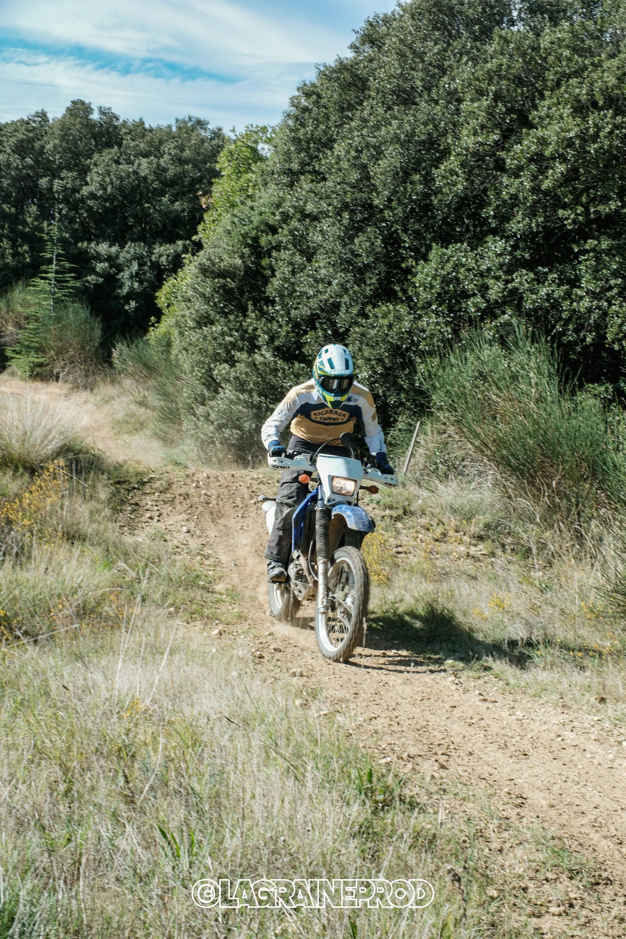 Un motocycliste portant un casque et des vêtements de protection chevauche une moto sur un sentier de terre en plein air, entouré de végétation et de buissons.