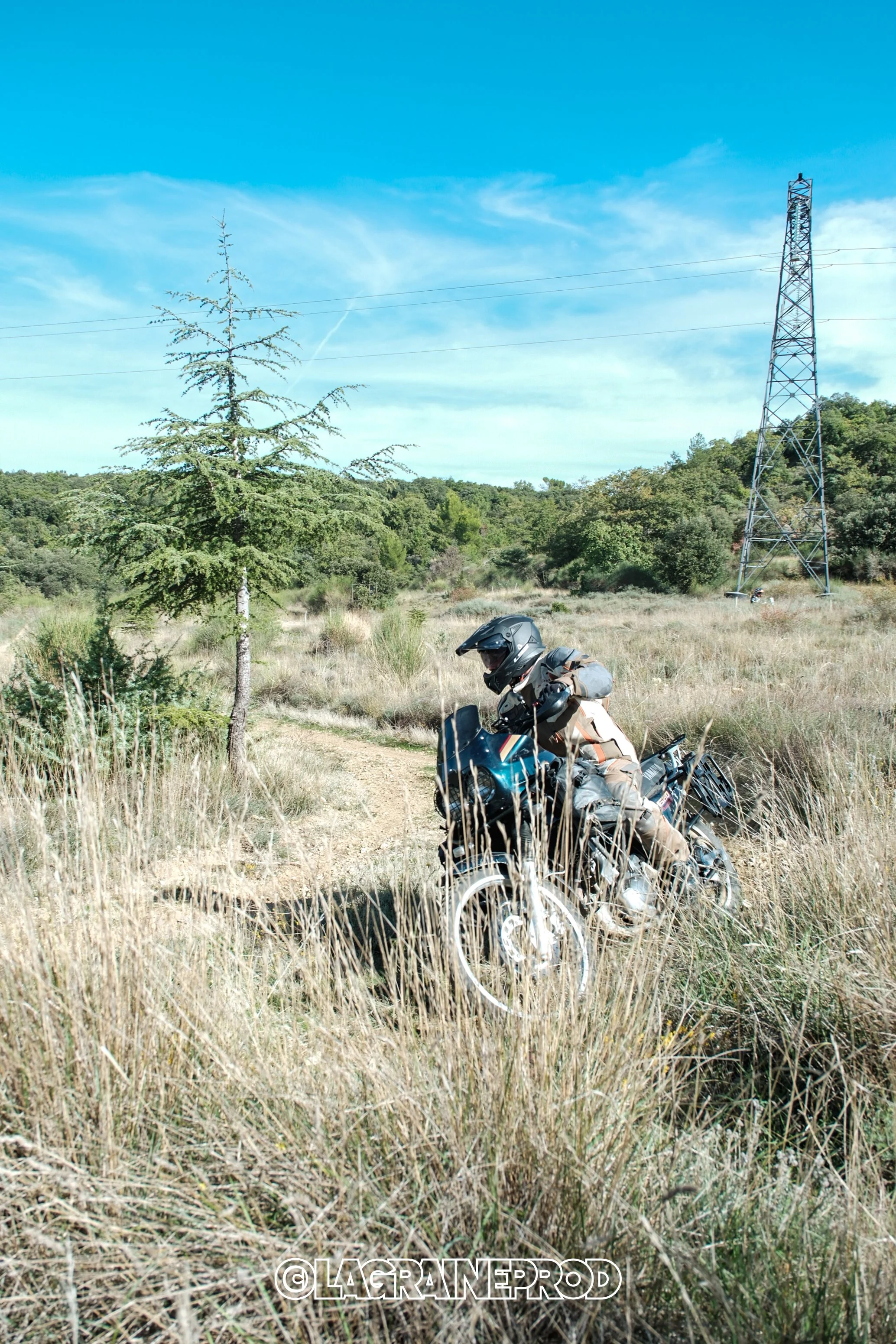 Un motard portant un casque, allongé sur une moto tout-terrain dans un paysage rural avec des herbes hautes, un arbre et une ligne électrique sous un ciel bleu avec quelques nuages.