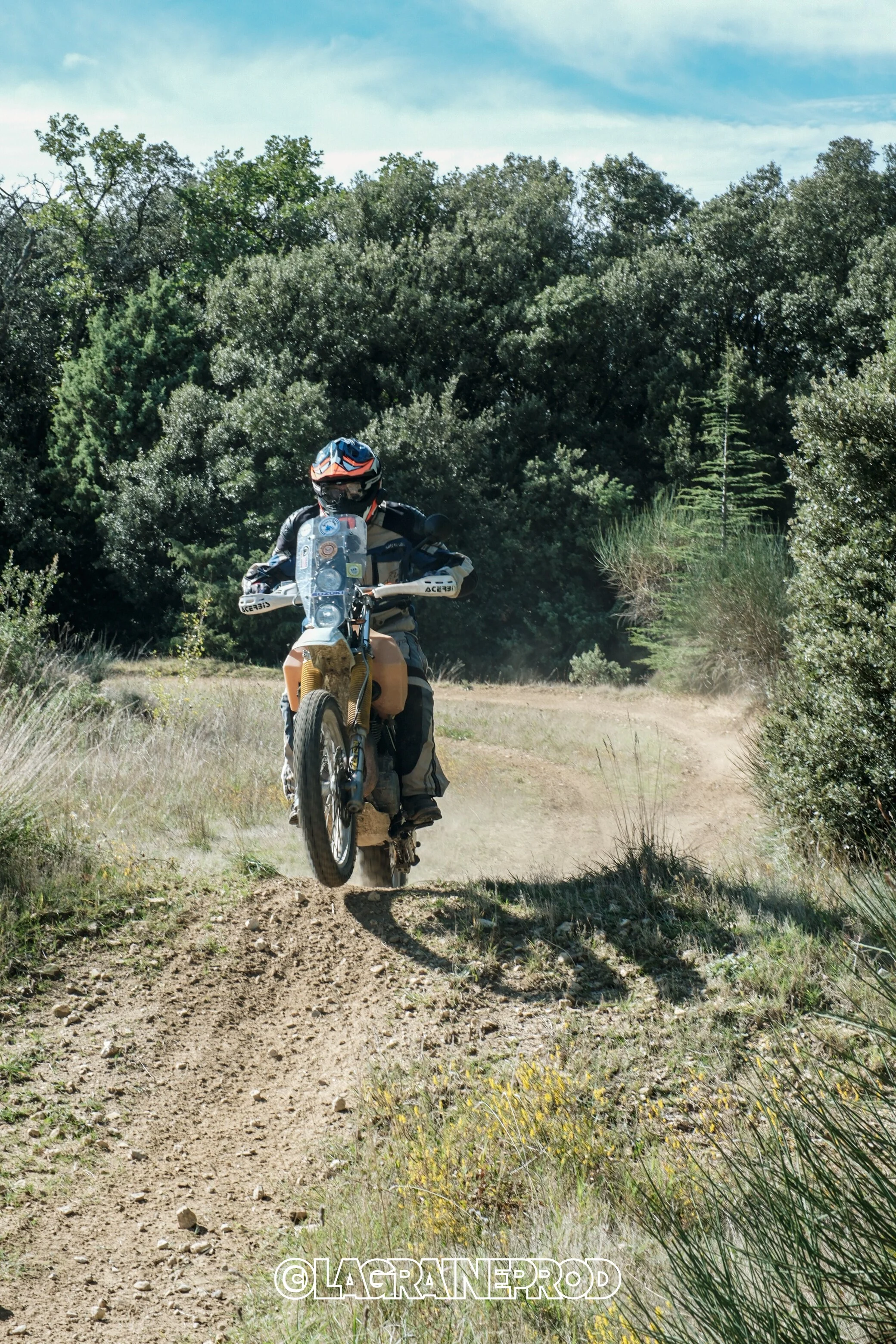 Un motocrosseur portant un casque et une combinaison, sautant un petit obstacle sur un sentier en plein air avec des arbres en arrière-plan.