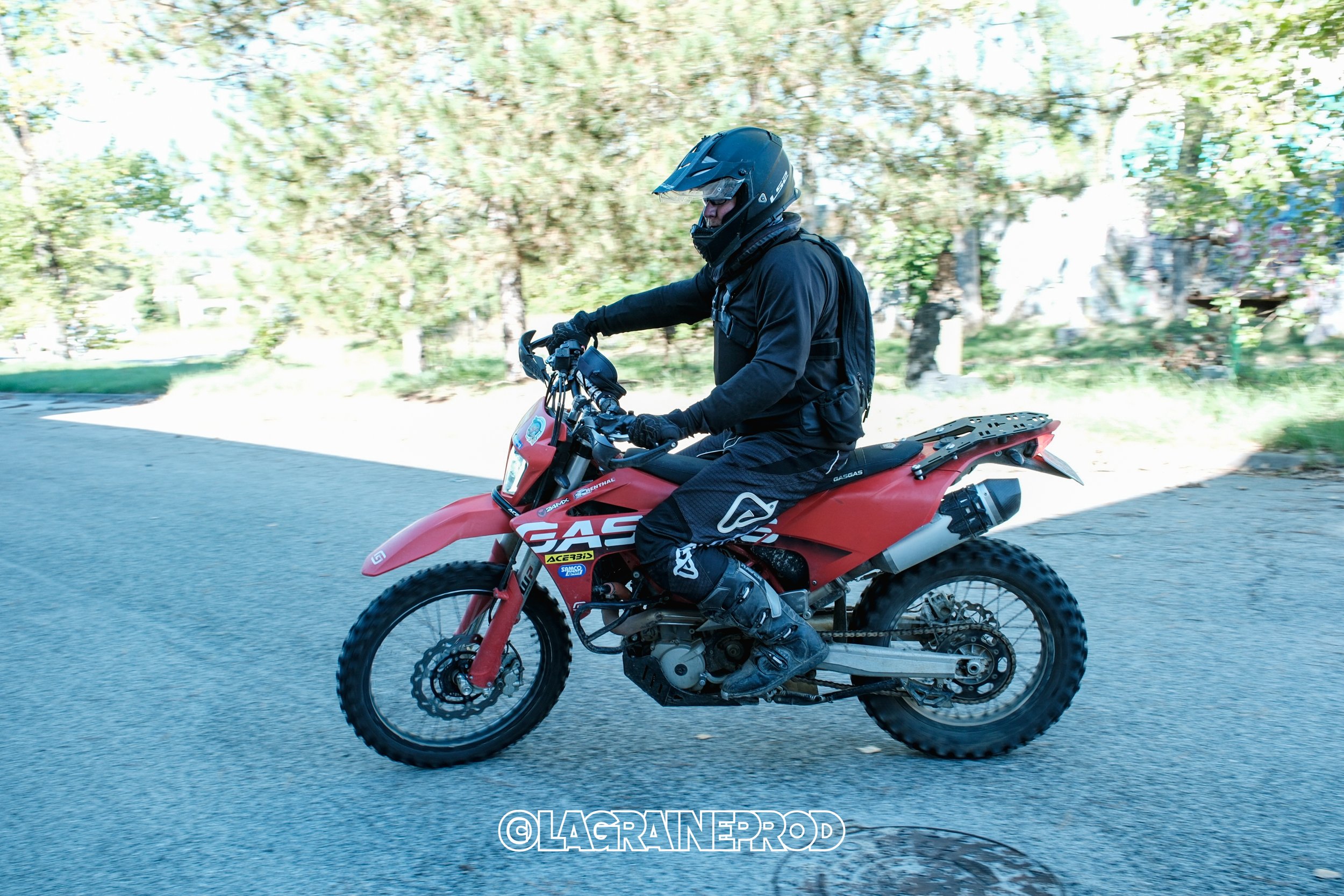 Un motocycliste en équipement noir roulant à moto rouge sur une route en zone boisée.