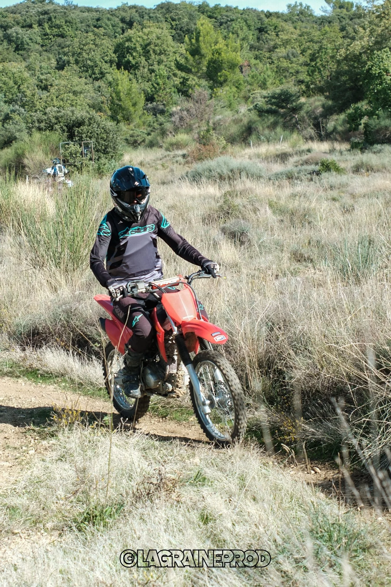 Jeune motocycliste portant un casque noir, une veste de motocross noire et un pantalon noir, chevauchant une moto rouge sur un sentier à travers un paysage rural avec de la végétation verte et des collines en arrière-plan.