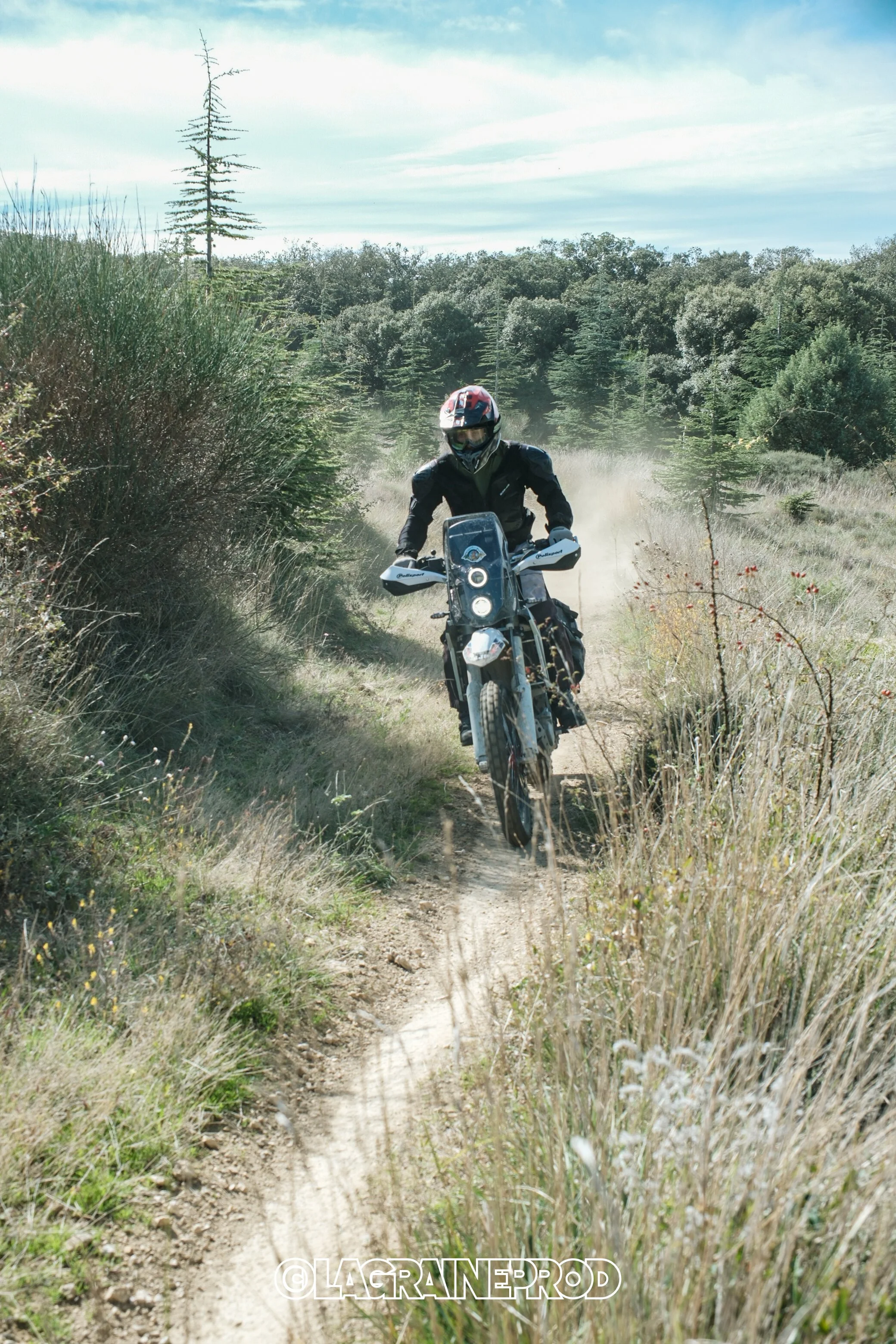 Un motard portant un casque roux, noir et blanc, circule sur un chemin de terre dans un environnement naturel avec des arbres et de l'herbe, créant de la poussière derrière lui.