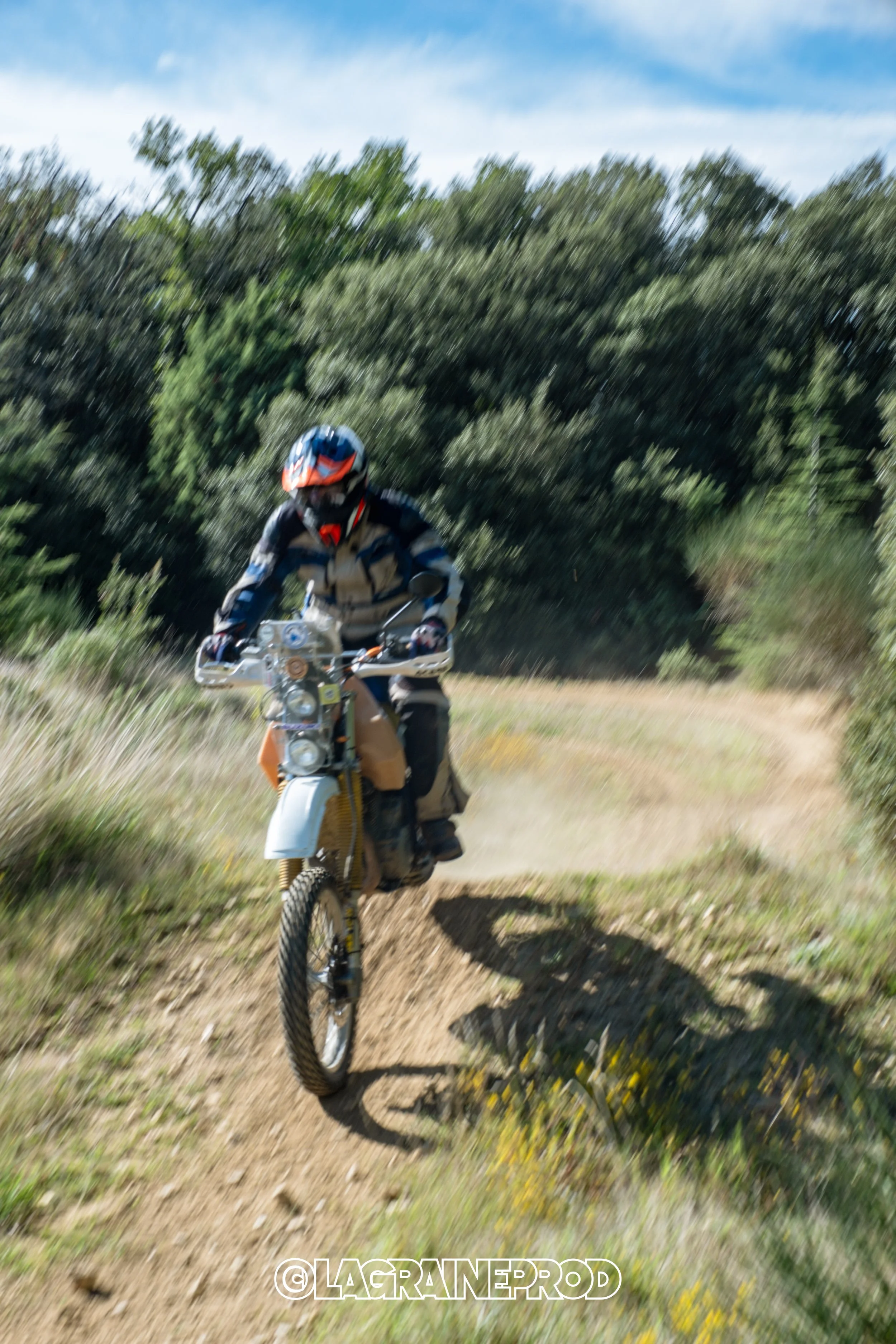 Coureur de motocross portant un casque et une tenue de protection, roulant sur un sentier en dehors de la route dans un paysage naturel avec des arbres en arrière-plan.