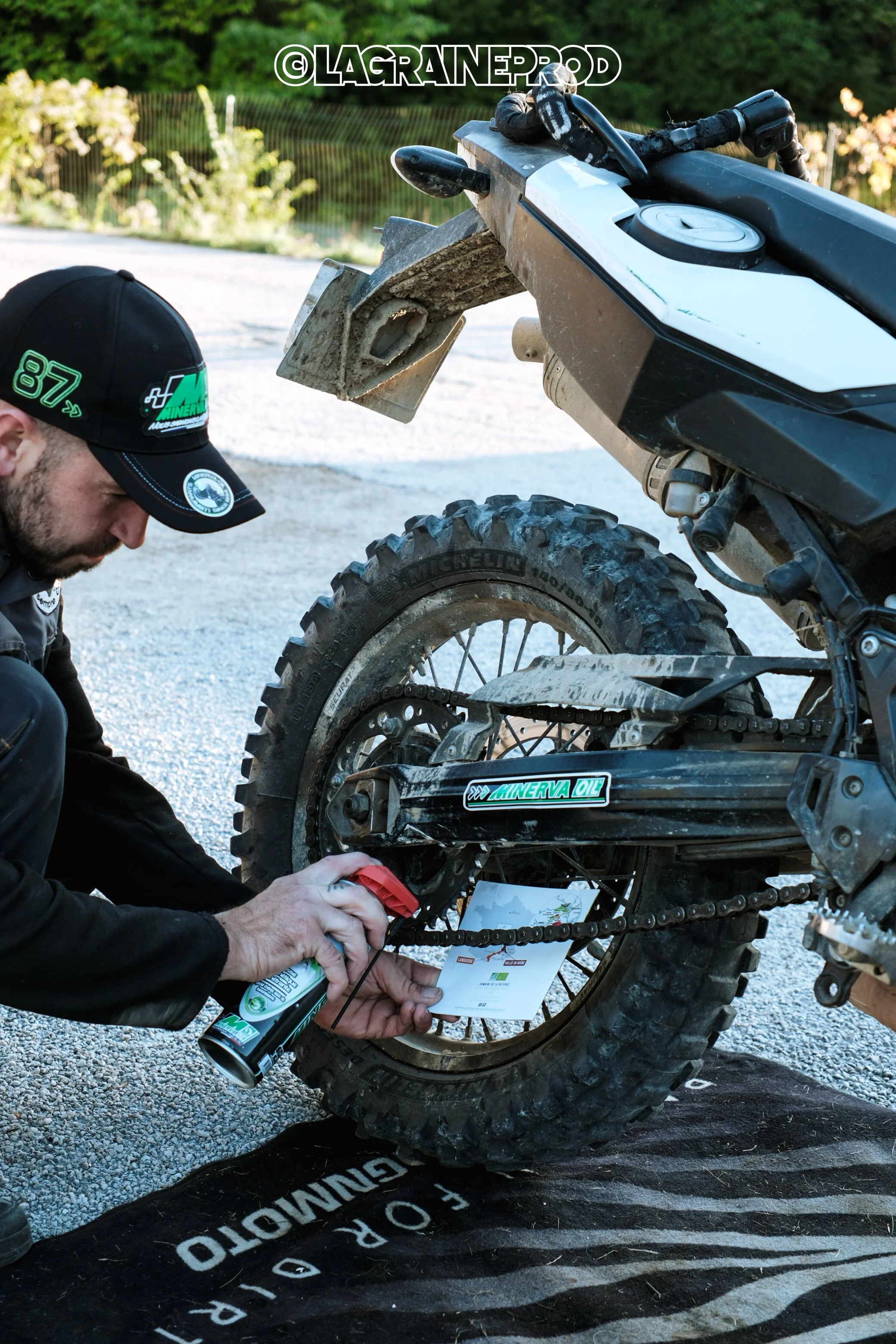 Un homme en train de graisser la chaîne d'une moto de dirt bike, avec un sol en gravier et des végétations en arrière-plan.