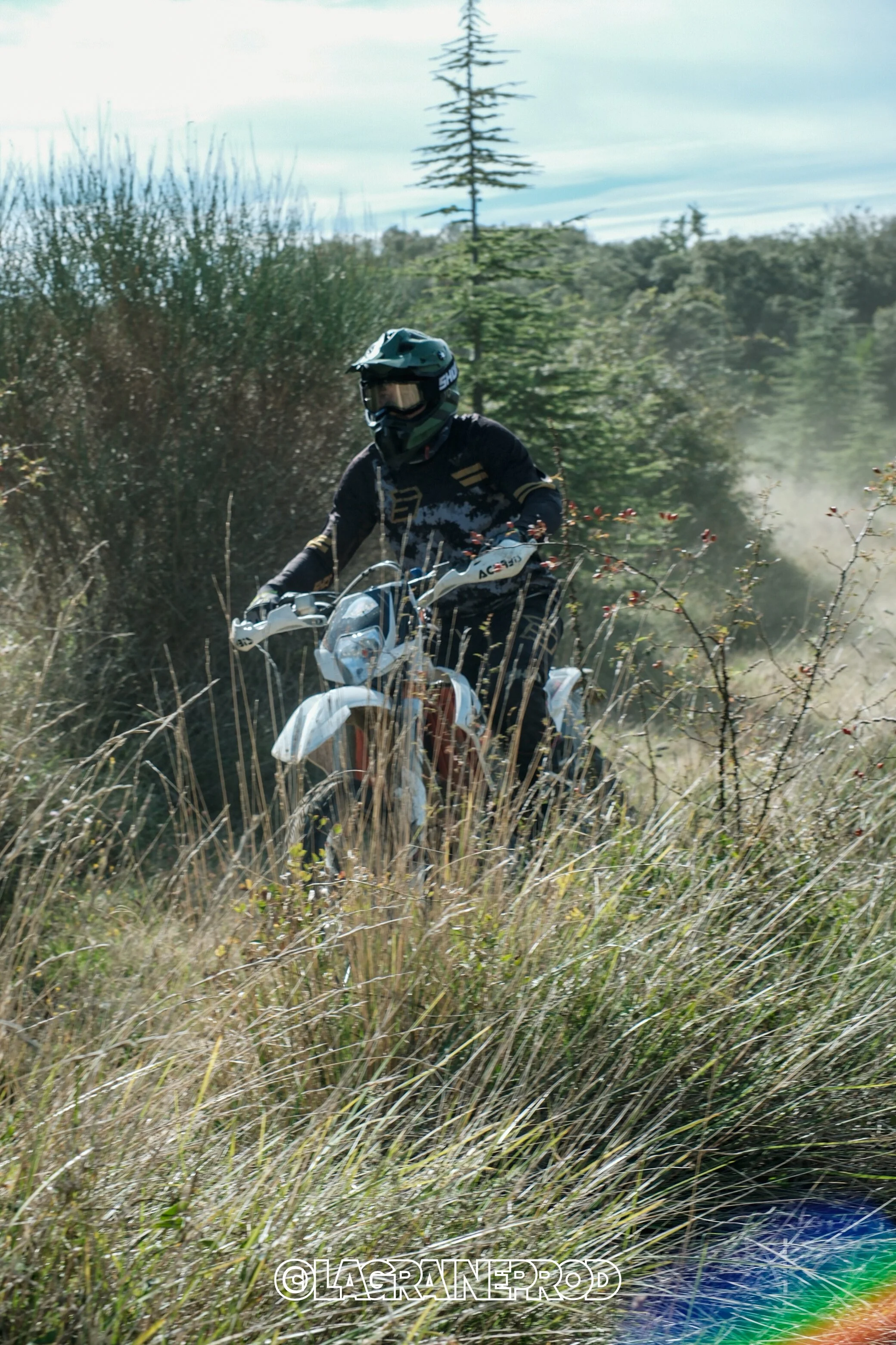 Une personne portant un casque de moto et un équipement de protection, chevauchant une moto tout-terrain à travers une zone de broussailles et d'herbes hautes dans un environnement naturel, avec des arbres en arrière-plan.