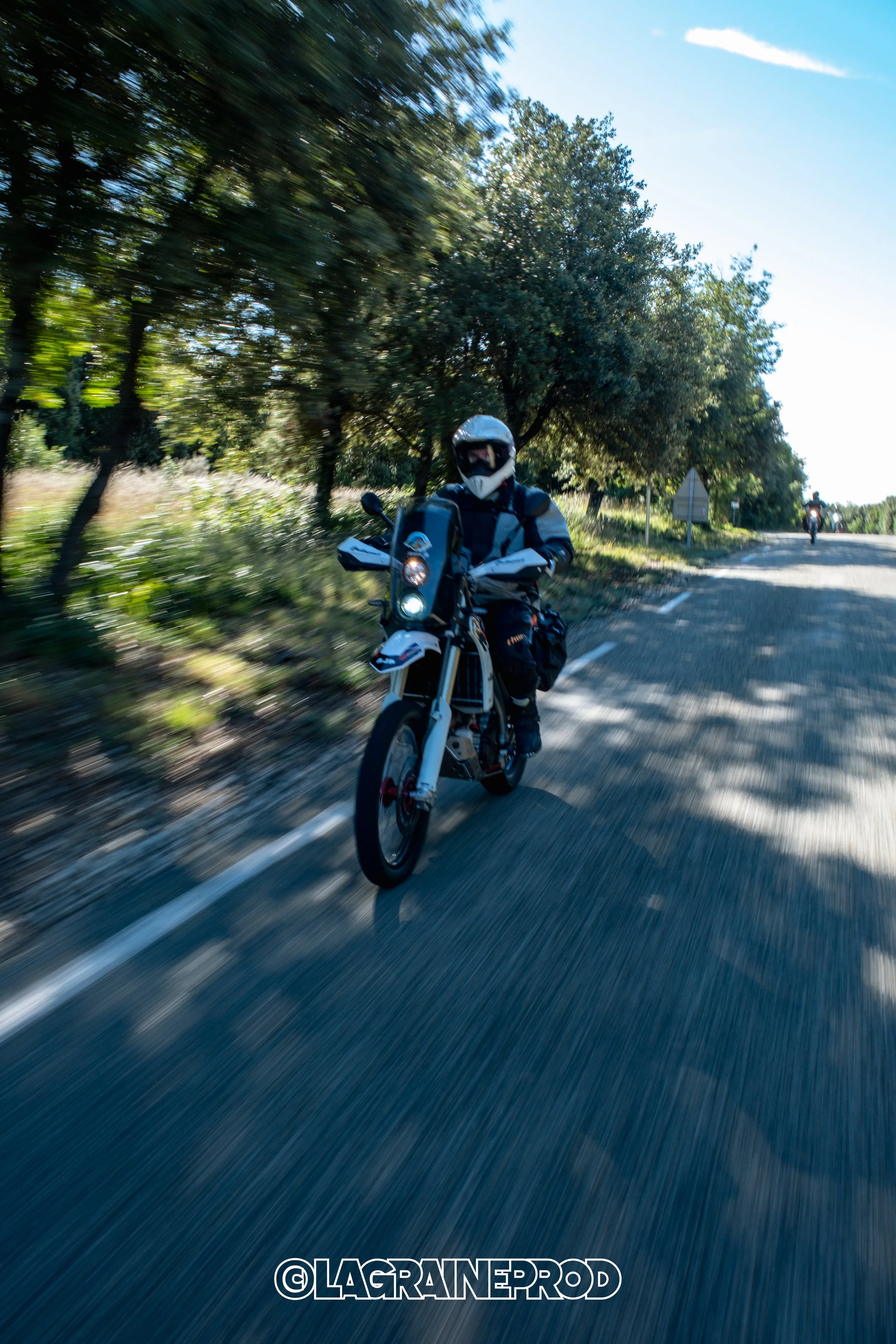 Motard portant un casque blanc et un équipement noir, roulant sur une route en campagne bordée d'arbres, avec un autre motocycliste visible en arrière-plan.