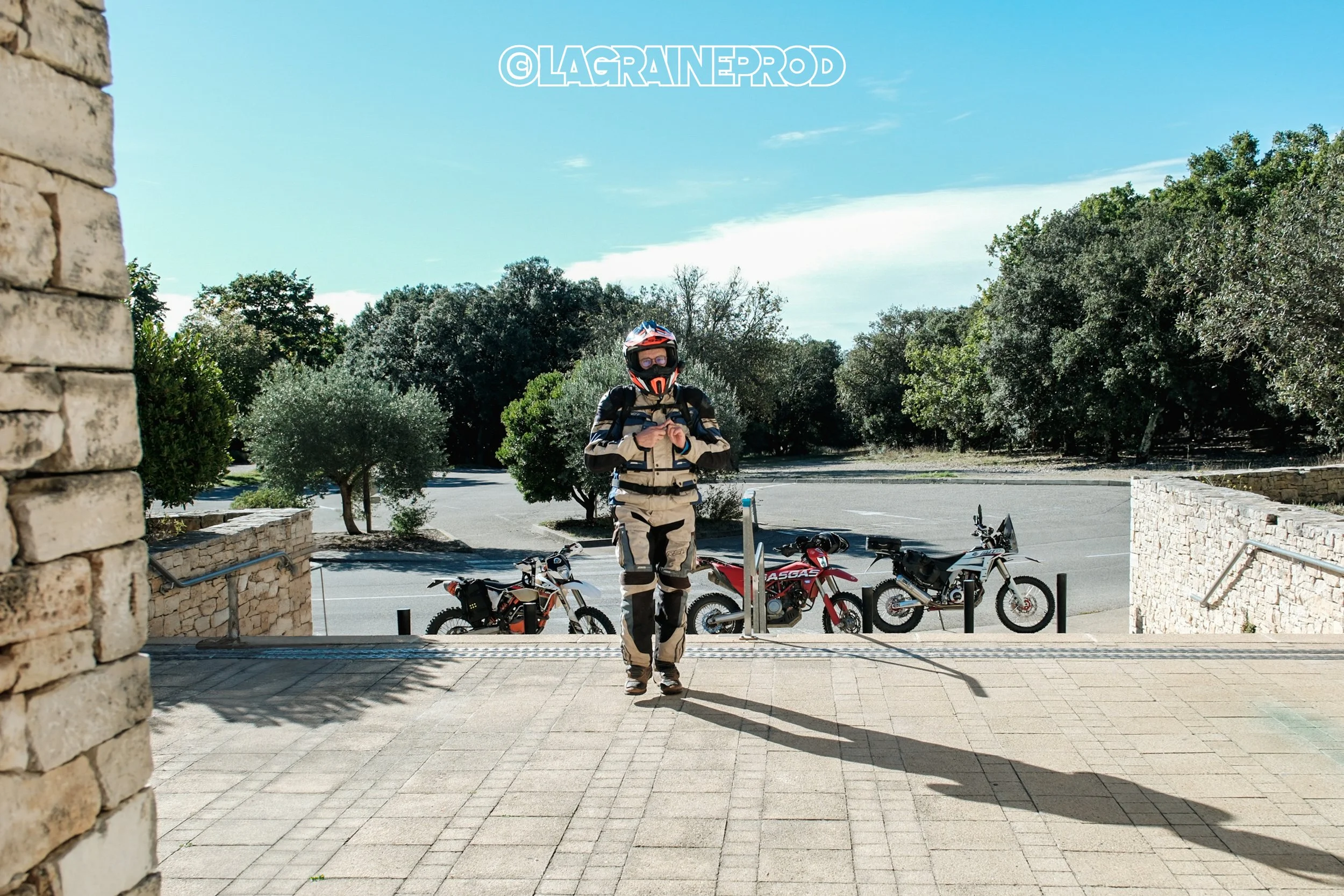 Une personne en tenue de moto tout-terrain avec un casque, debout en face de trois vélos électriques, à l'entrée d'une zone piétonne bordée d'arbres, sous un ciel bleu clair.