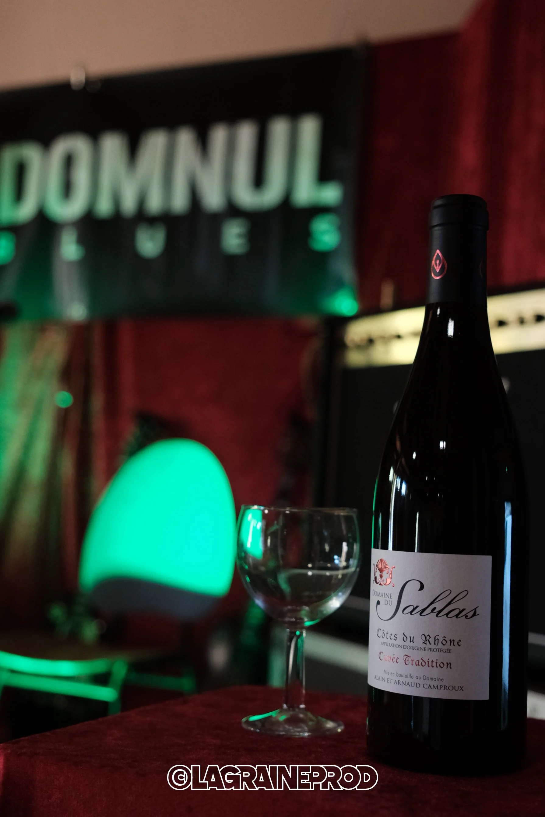 Une bouteille de vin Rouge, un verre à vin vide, une lampe verte et un fond avec un panneau 'DOMINUL' en lettres lumineuses. 