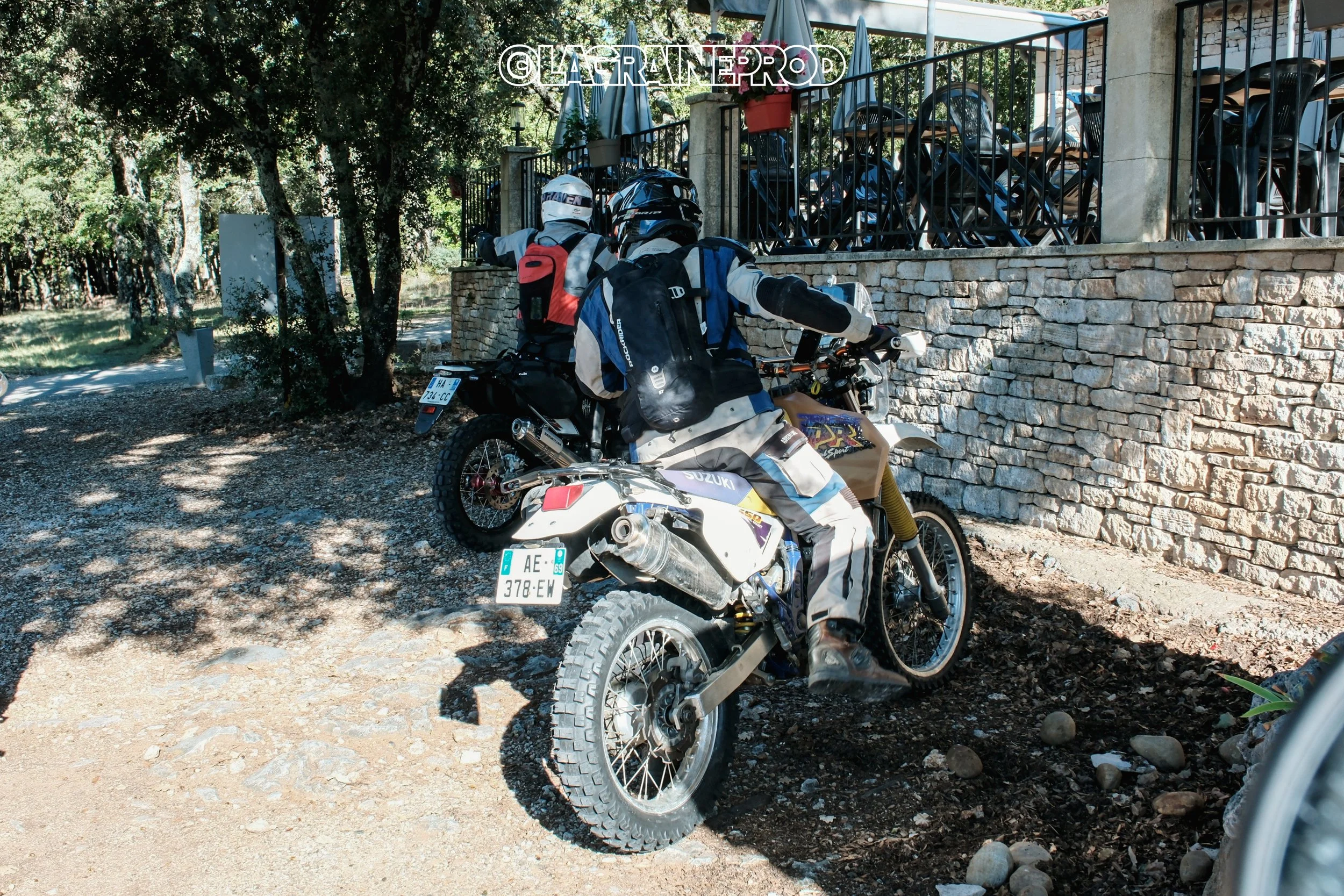 Deux motards en tenue de protection, avec casques, s'apprêtent à partir à moto sur un chemin herbeux à proximité d'une clôture en pierre, avec des arbres et un parking à motos en arrière-plan.