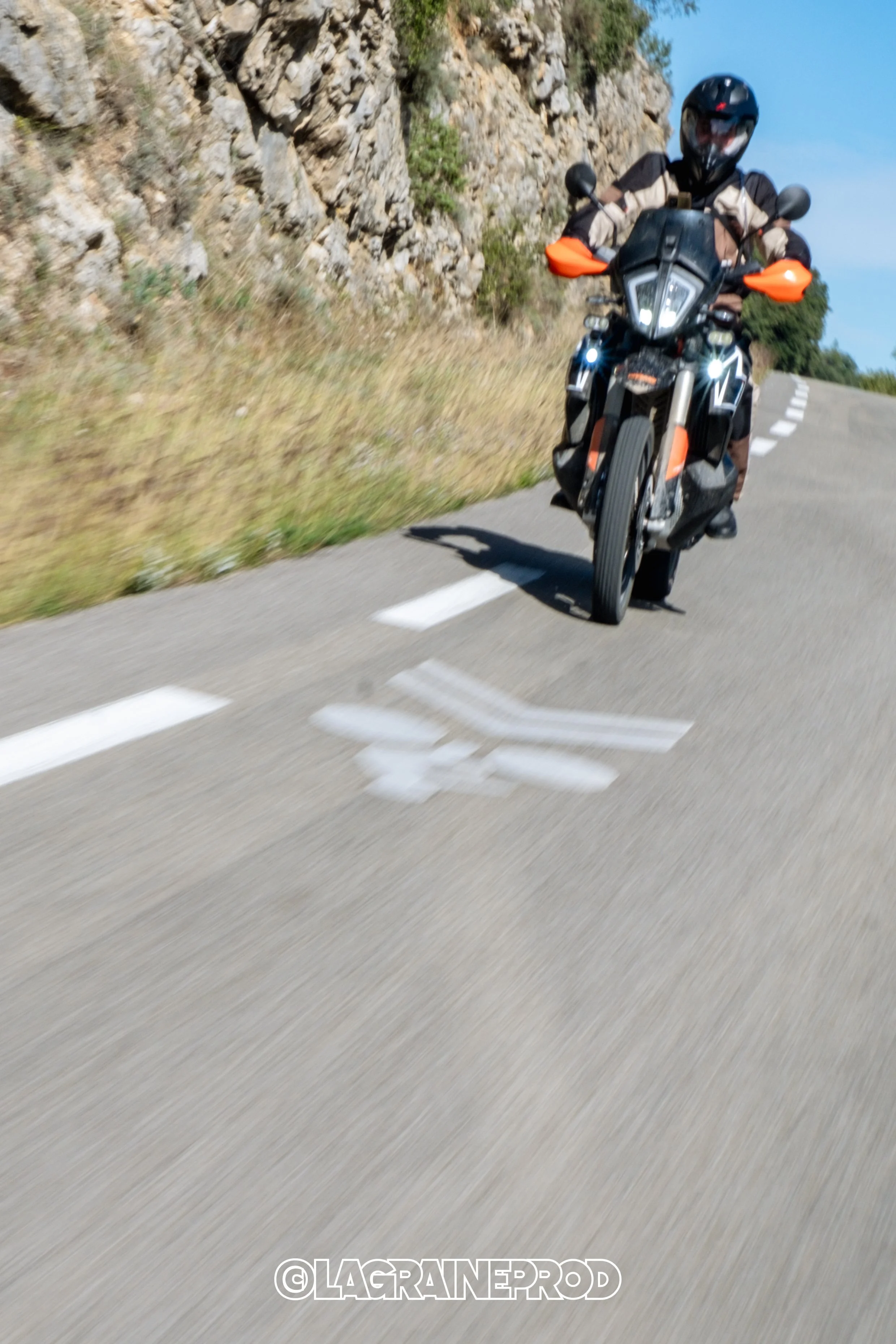 Un motard portant un casque noir, une veste noire et beige, roulant sur une route sinueuse avec un panneau blanc au sol et des rochers le long du bord de la route, ciel bleu en arrière-plan.
