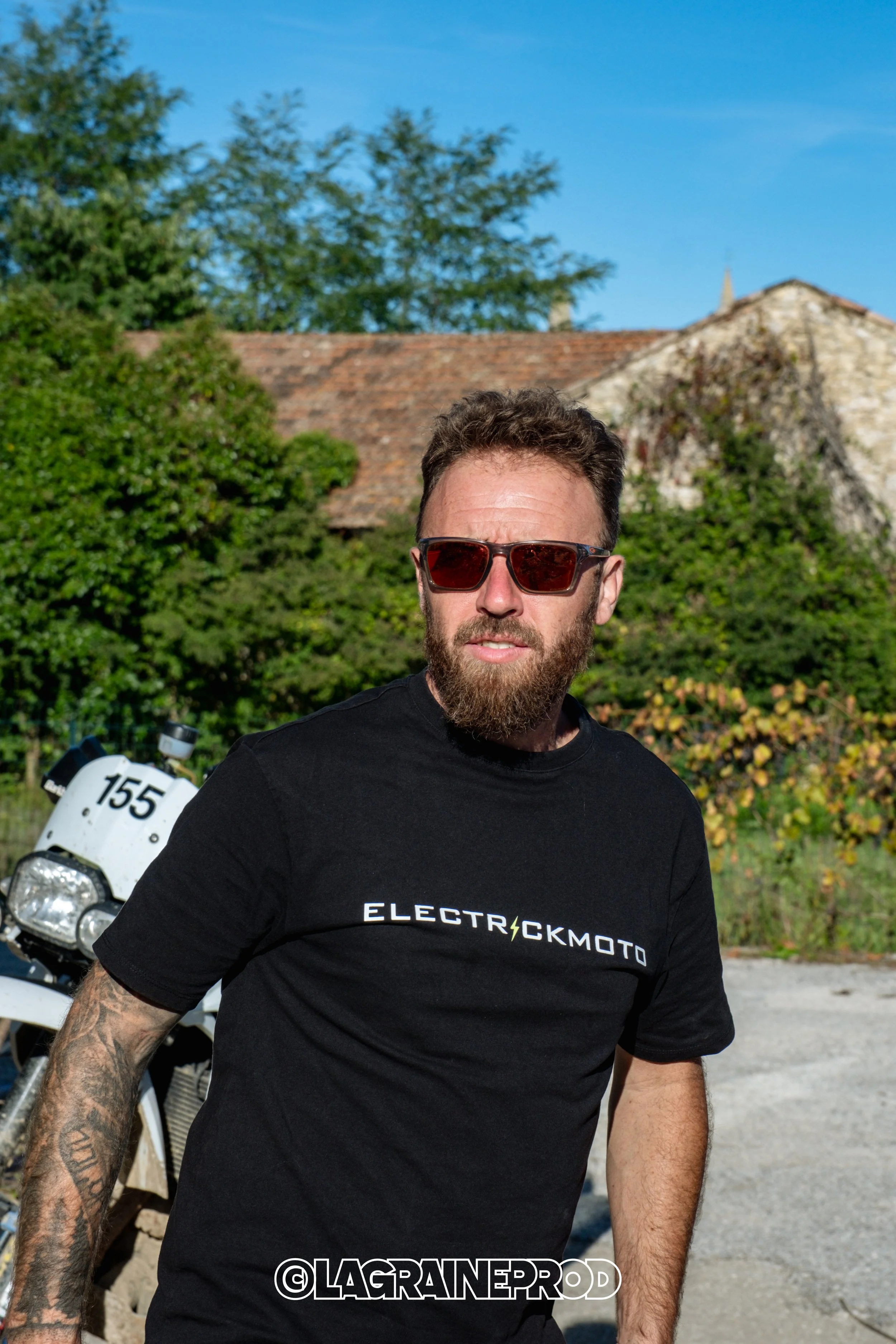 Un homme avec une barbe portant des lunettes de soleil, un t-shirt noir 'ELECTRICKMOTO', en extérieur avec un vélo dans le fond et une maison en pierre derrière lui, sous un ciel bleu.