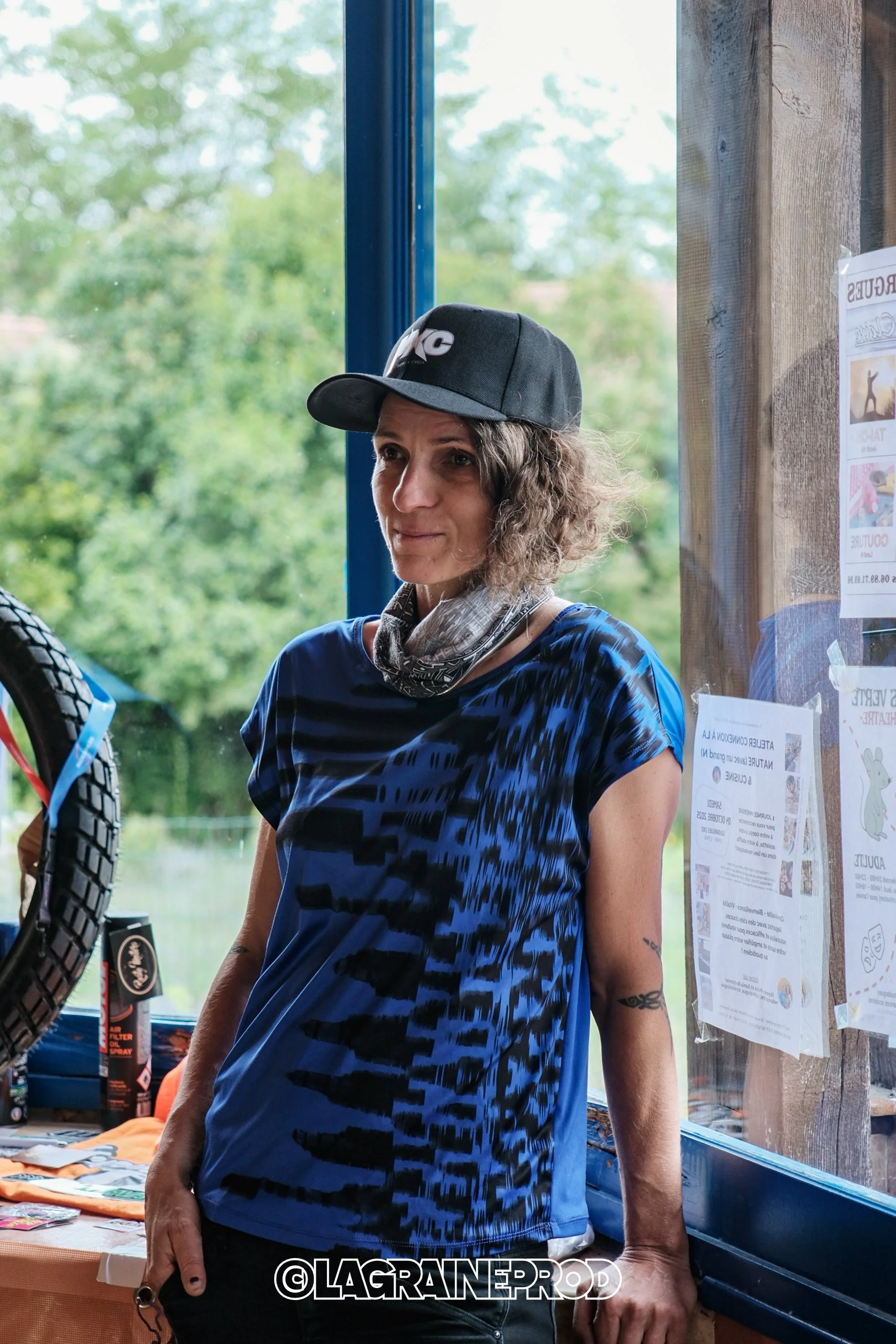 Une femme avec une casquette noire, une chemise bleue à motif, et un foulard autour du cou, se tient à l'intérieur près d'une fenêtre, avec un arrière-plan de nature verte.