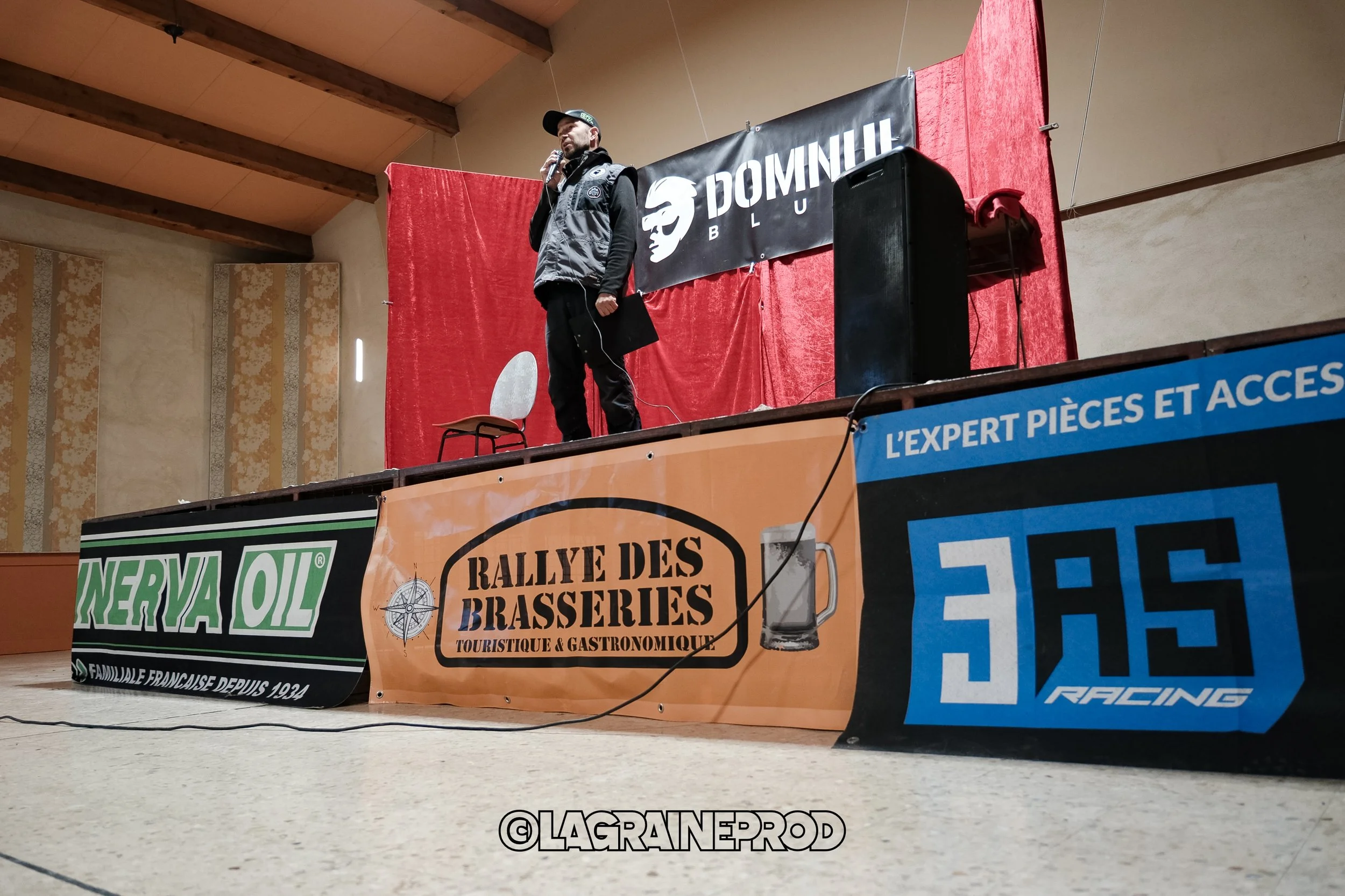 Un homme en veste et casquette parle lors d'une présentation ou d'une conférence sur scène, derrière un fond rouge avec une bannière 'DOMINILH'. La scène a des banderoles publicitaires, dont une pour 'Rallye des Brasseries' et une autre pour 'Nerya O