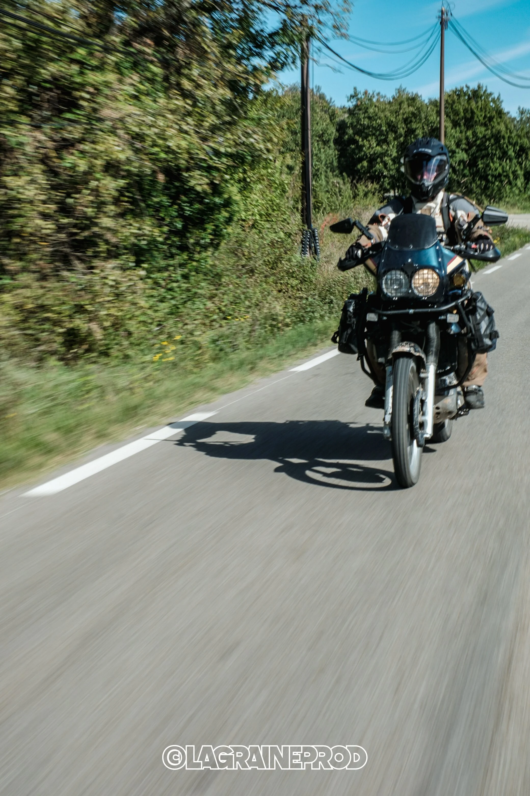 Un motard portant un casque noir, roulant sur une route de campagne bordée d'arbres, avec des poteaux électriques à côté.