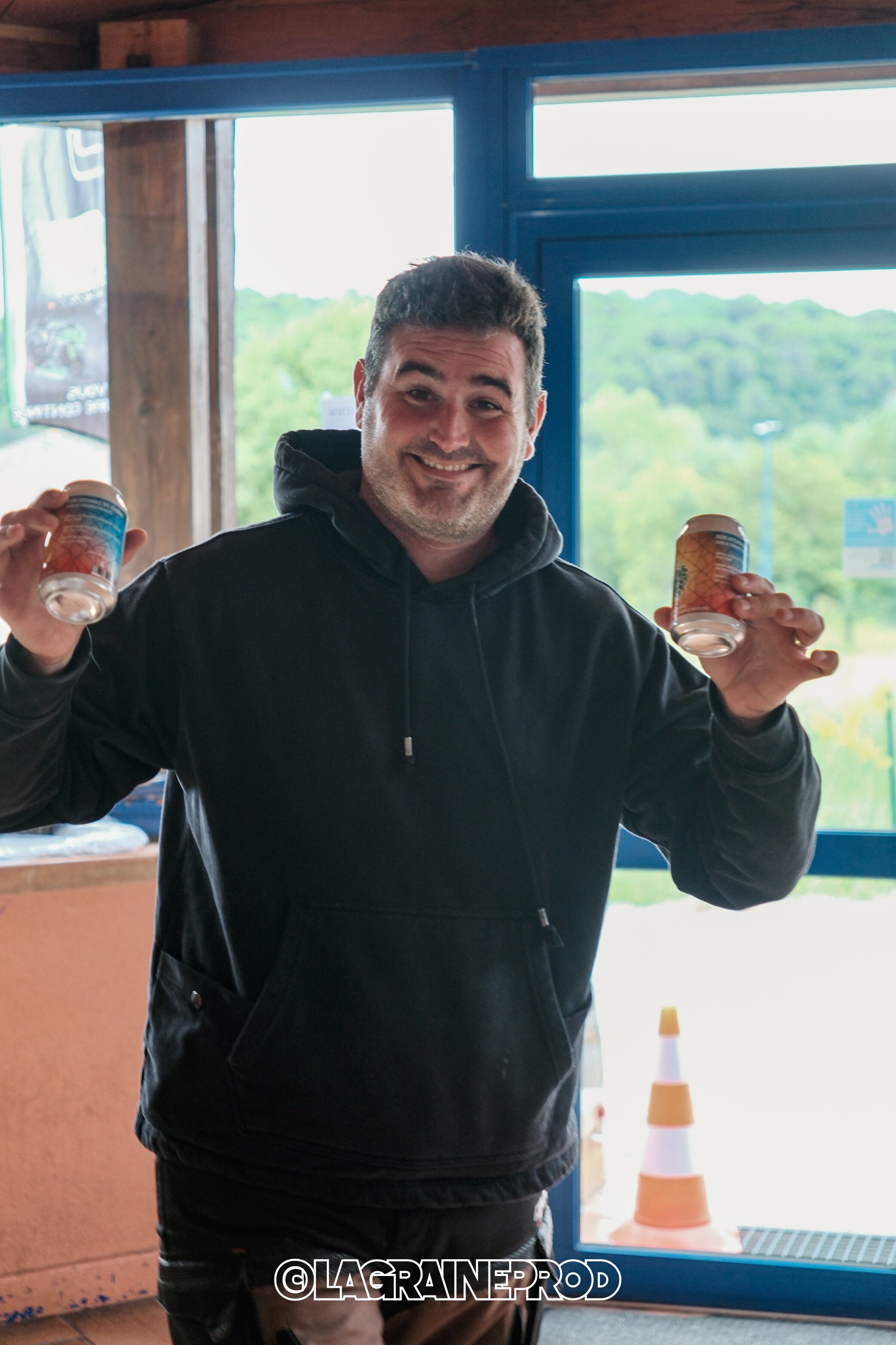 Un homme souriant tient deux canettes, une dans chaque main, à l'intérieur d'un bâtiment avec une grande fenêtre laissant voir un paysage vert en arrière-plan.