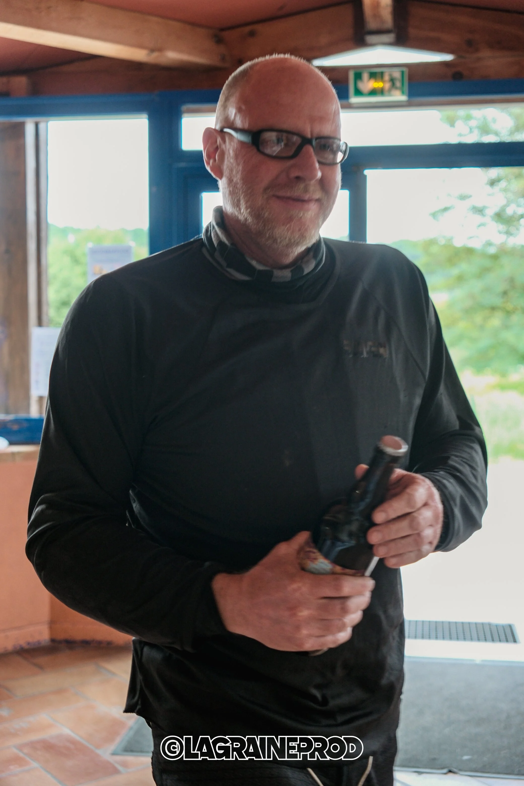 Un homme chauve avec des lunettes, tenant une bouteille de bière, à l'intérieur d'un bâtiment en bois avec grande porte vitrée et vue sur la nature en arrière-plan.