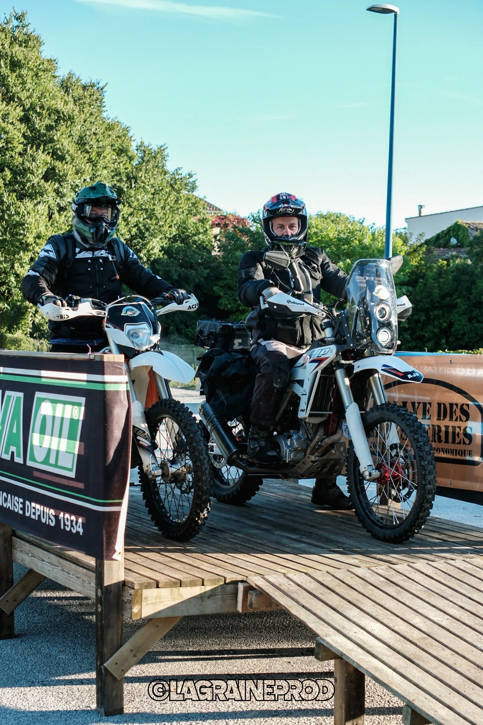 Deux motards portant des casques et des vestes en cuir, posant avec leurs motos tout-terrain sur une plateforme en bois, à l'extérieur, avec des arbres et un lampadaire en arrière-plan.