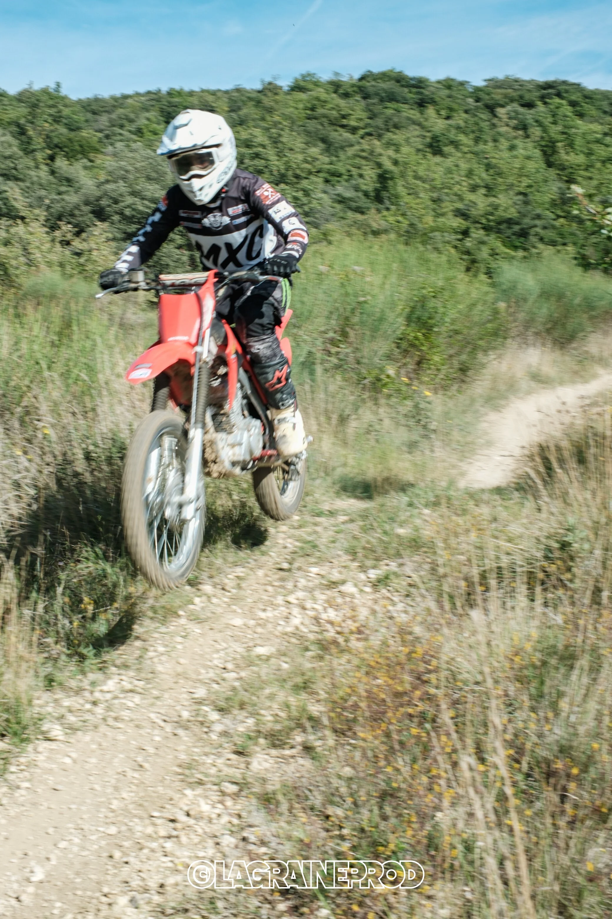 Motocross en plein air sur un sentier de terre, pilote portant un casque blanc et une tenue de moto, dans un paysage verdoyant avec des collines en arrière-plan.