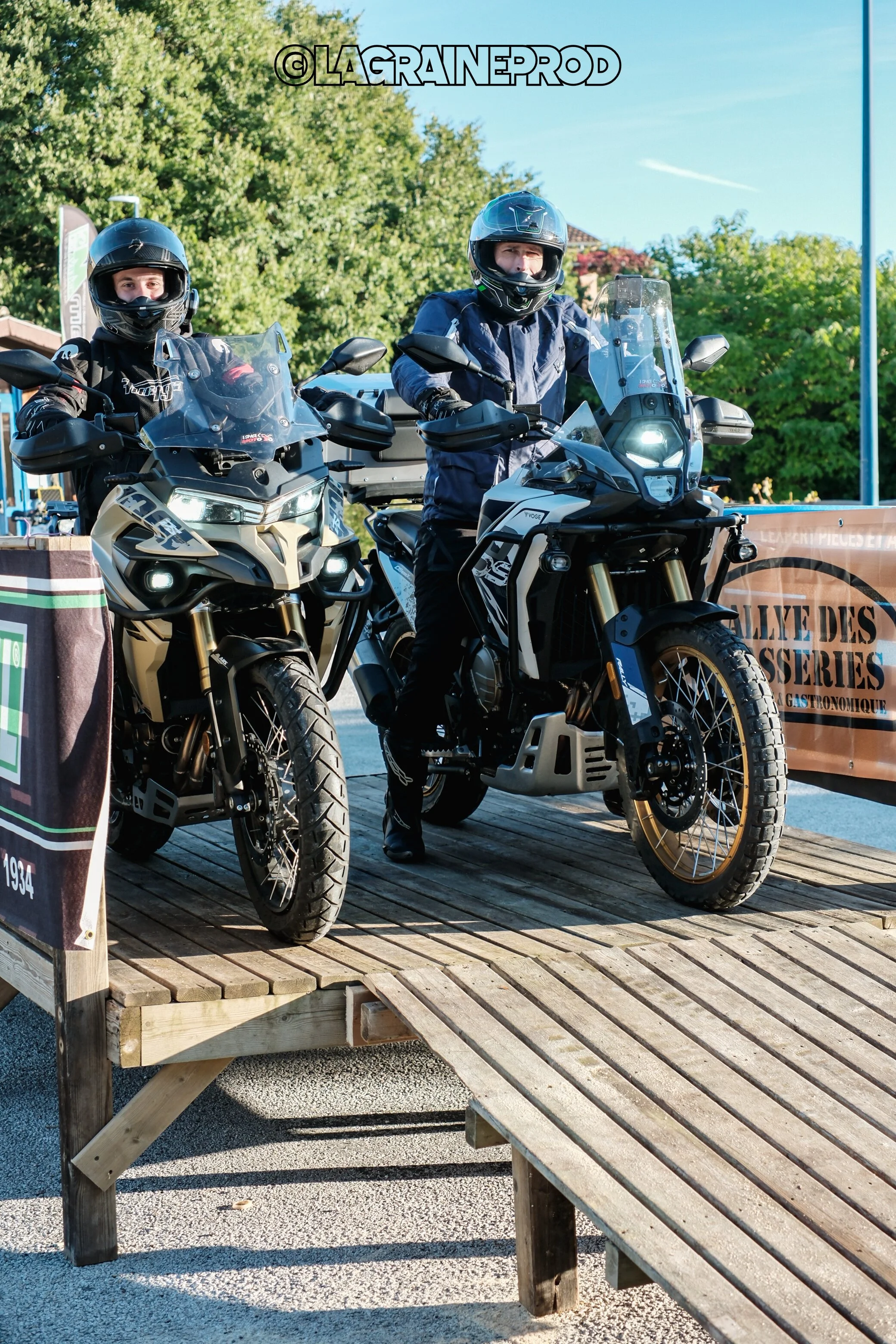 Deux motocyclistes en tenue de protection sur des motos tout-terrain, se tenant sur un pont en bois, avec un environnement extérieur vert et un ciel bleu.